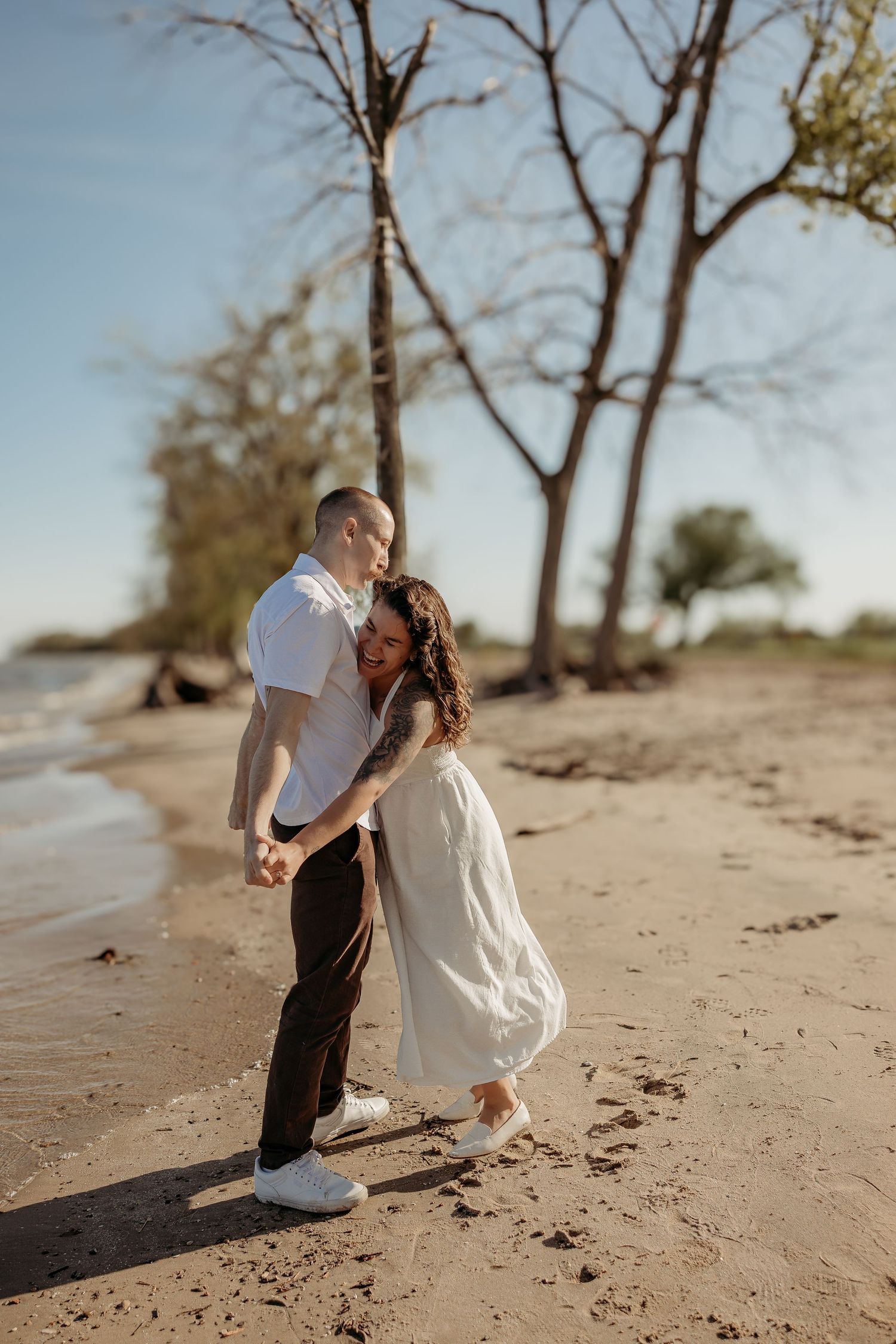 A couple embraces on a sandy beach at sunset, with bare trees creating a romantic silhouette in the background.