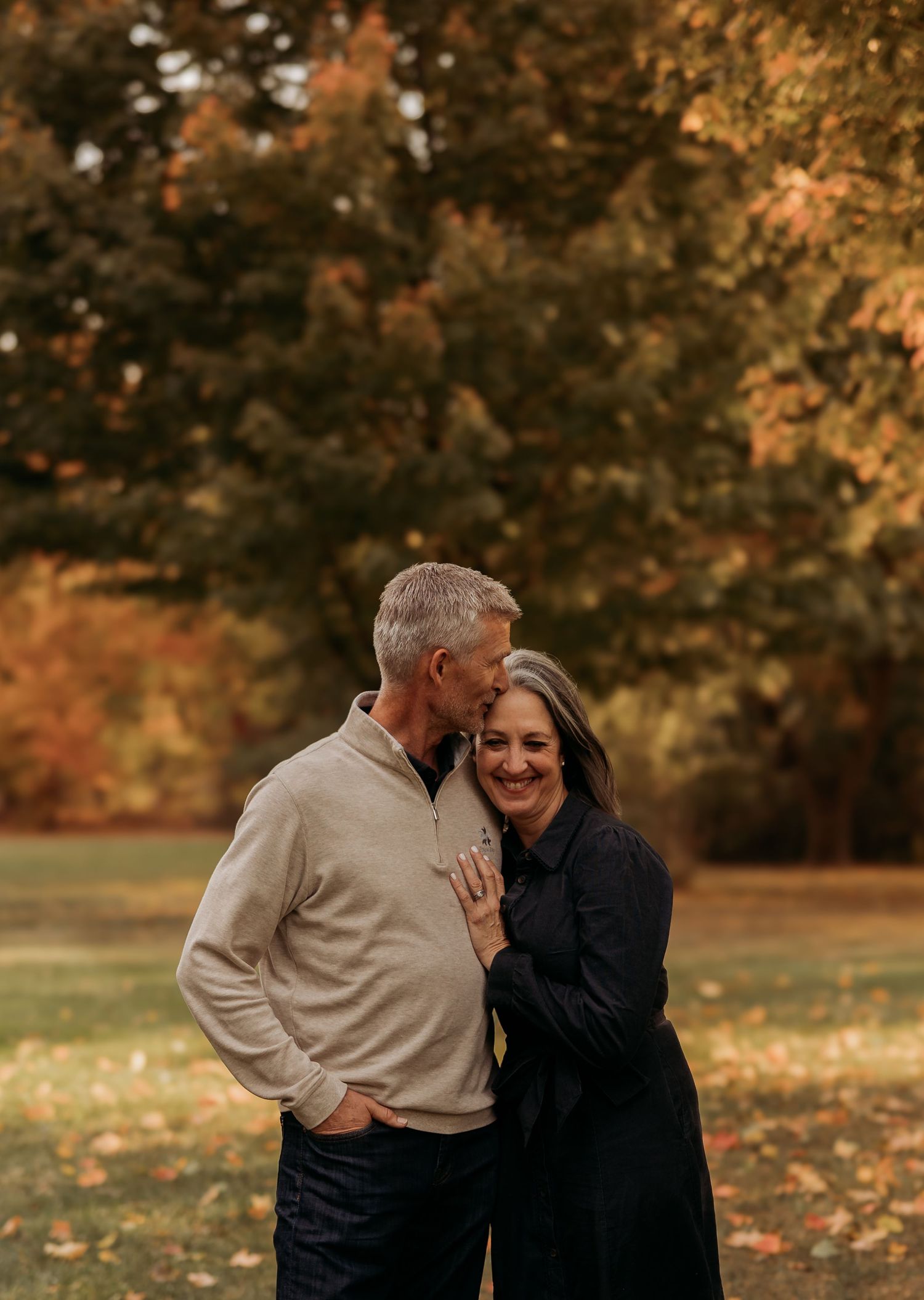 A couple shares tender moments in a park during autumn with golden fall foliage in the background.