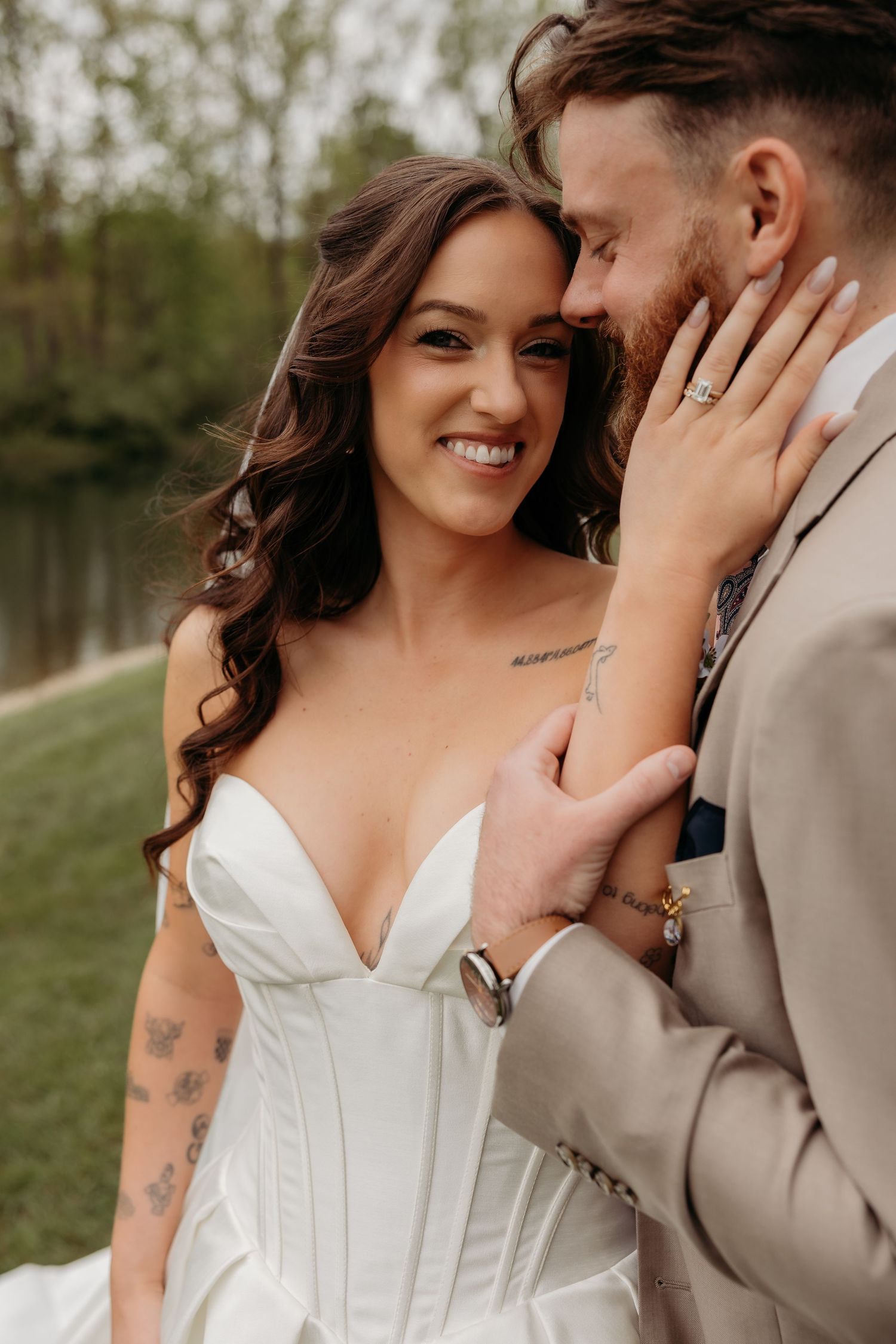 Smiling bride in white corset gown with tattoos poses with groom in tan suit outdoors near a lake.