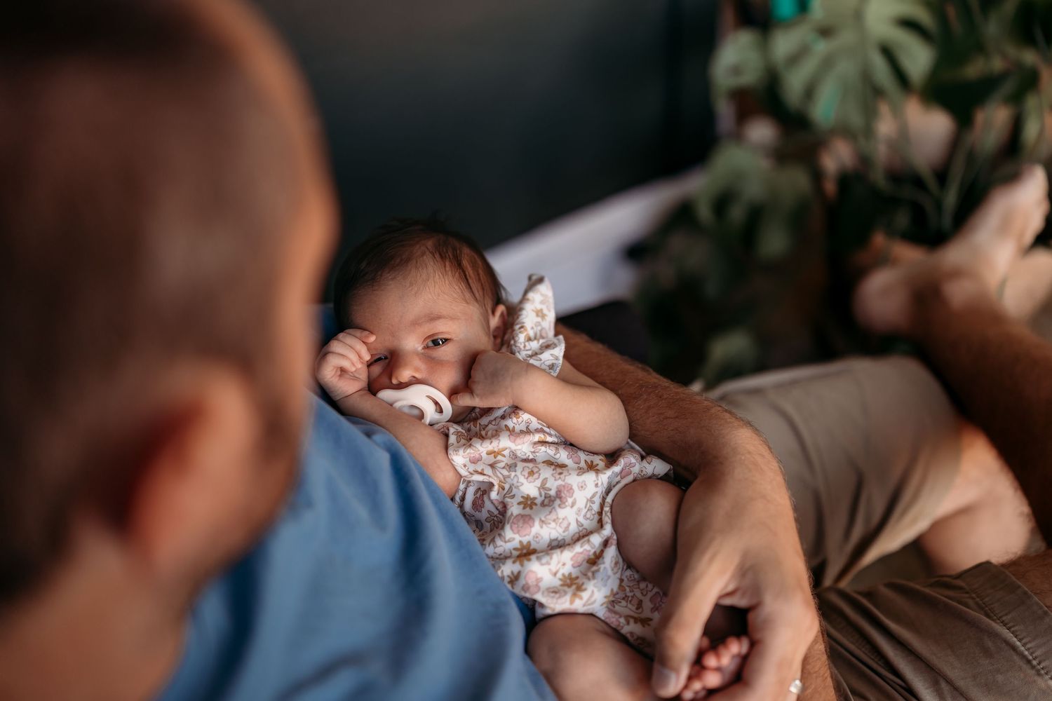 A sleeping infant wearing a floral romper lies peacefully in someone's arms while being admired.