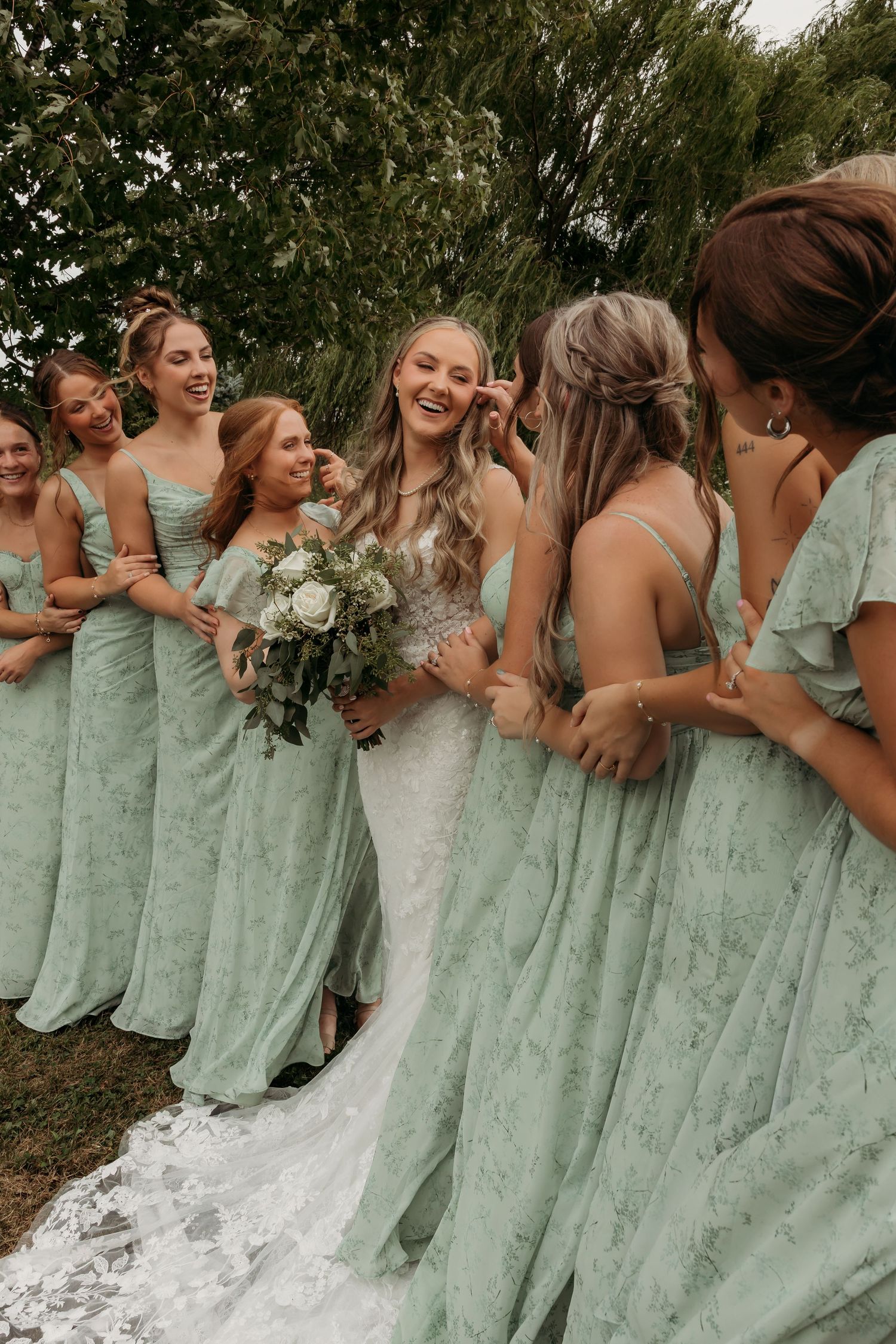 Bridesmaids in sage green dresses and bride in white gown pose together for a candid group photo outdoors.