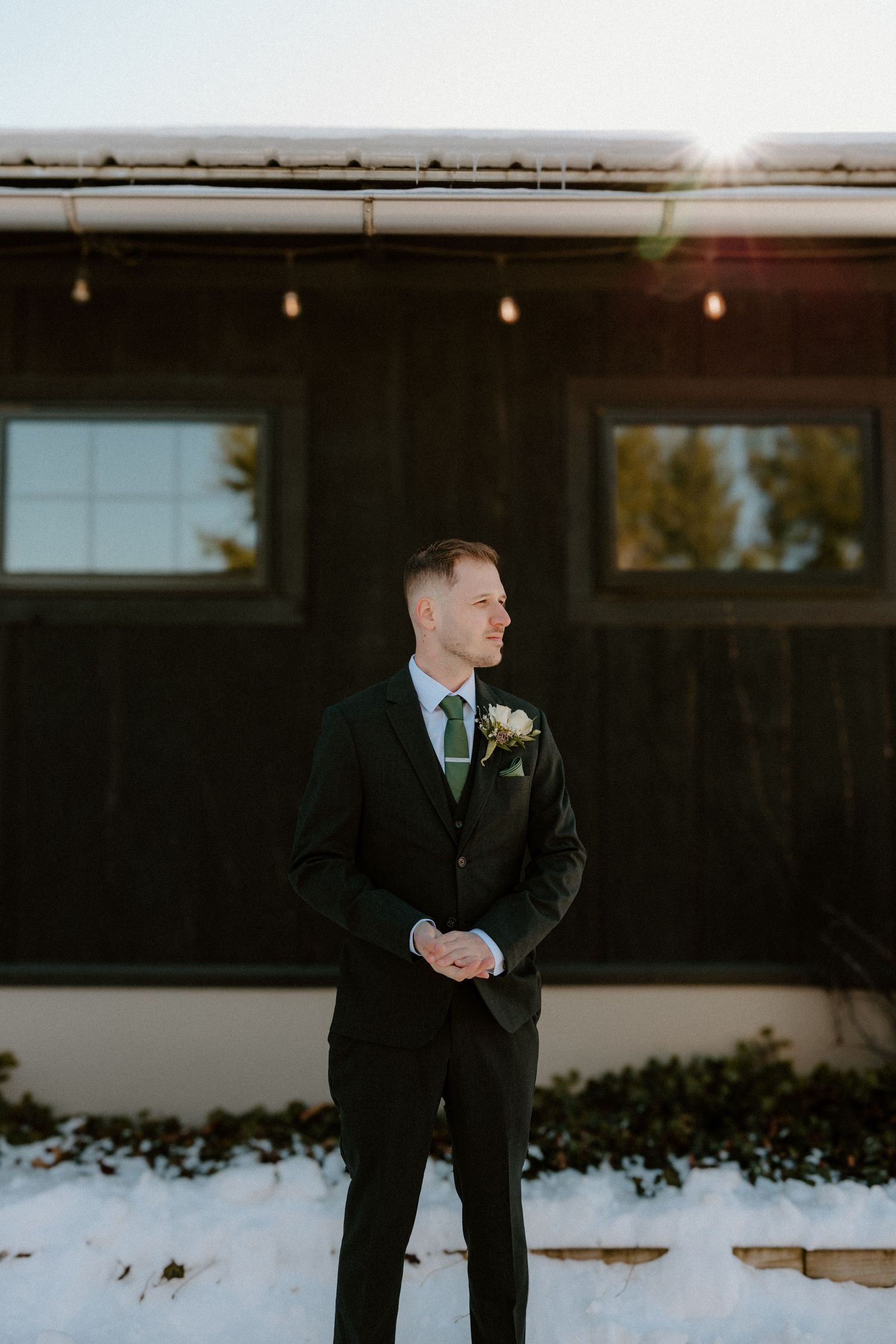 A groom looks pensive while standing outside in a black suit among snowy winter scenery at dusk.