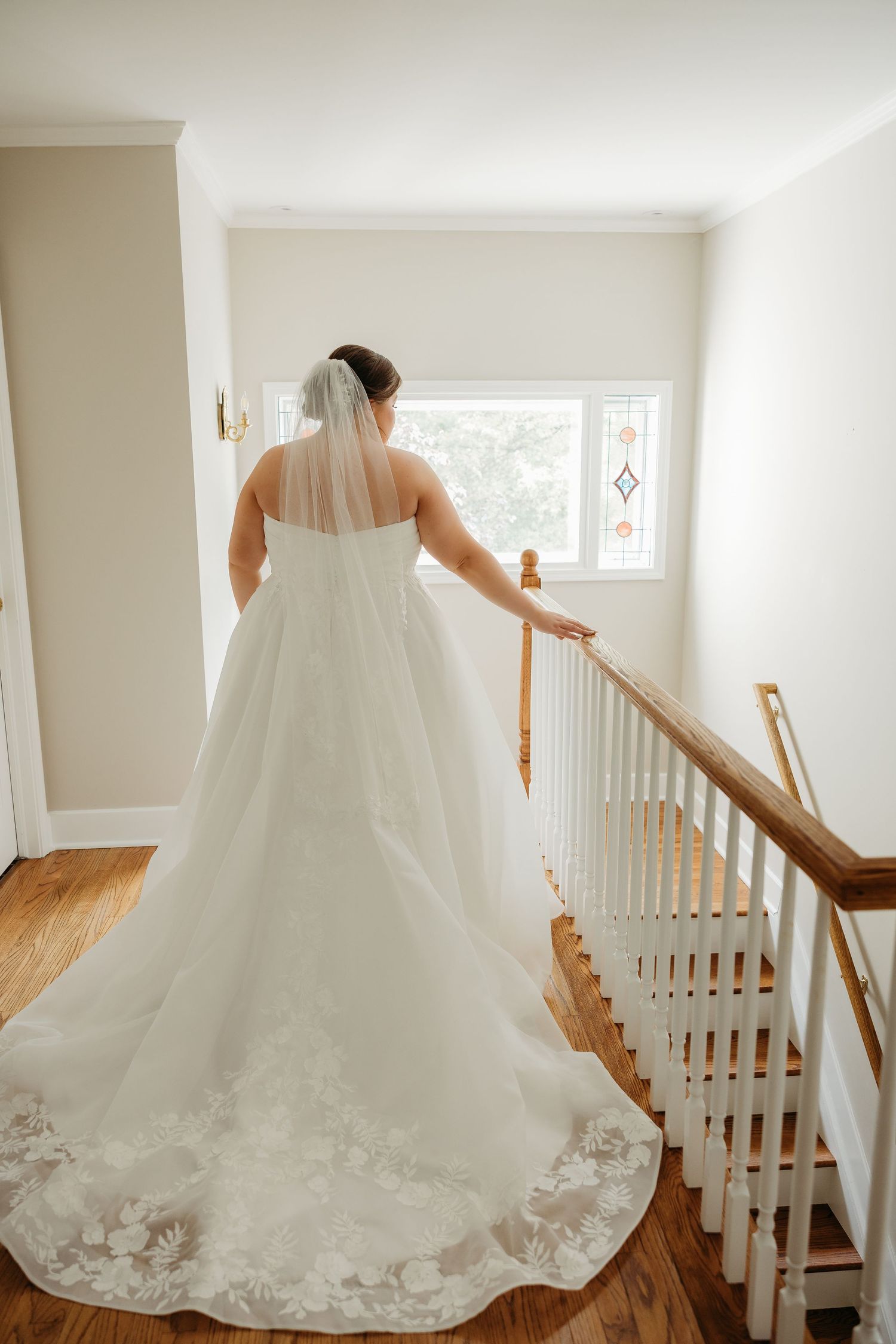 Bride in a white lace ball gown with veil stands at staircase railing, viewed from behind near window.