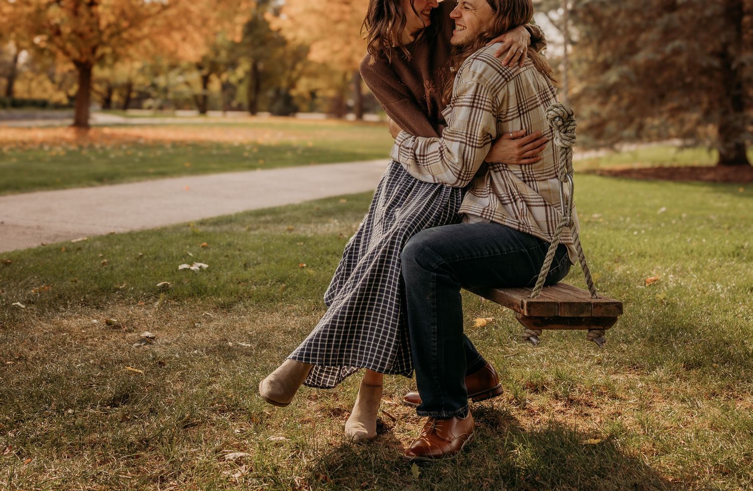 A couple in fall attire sits together on a rustic wooden swing in an autumn park setting.