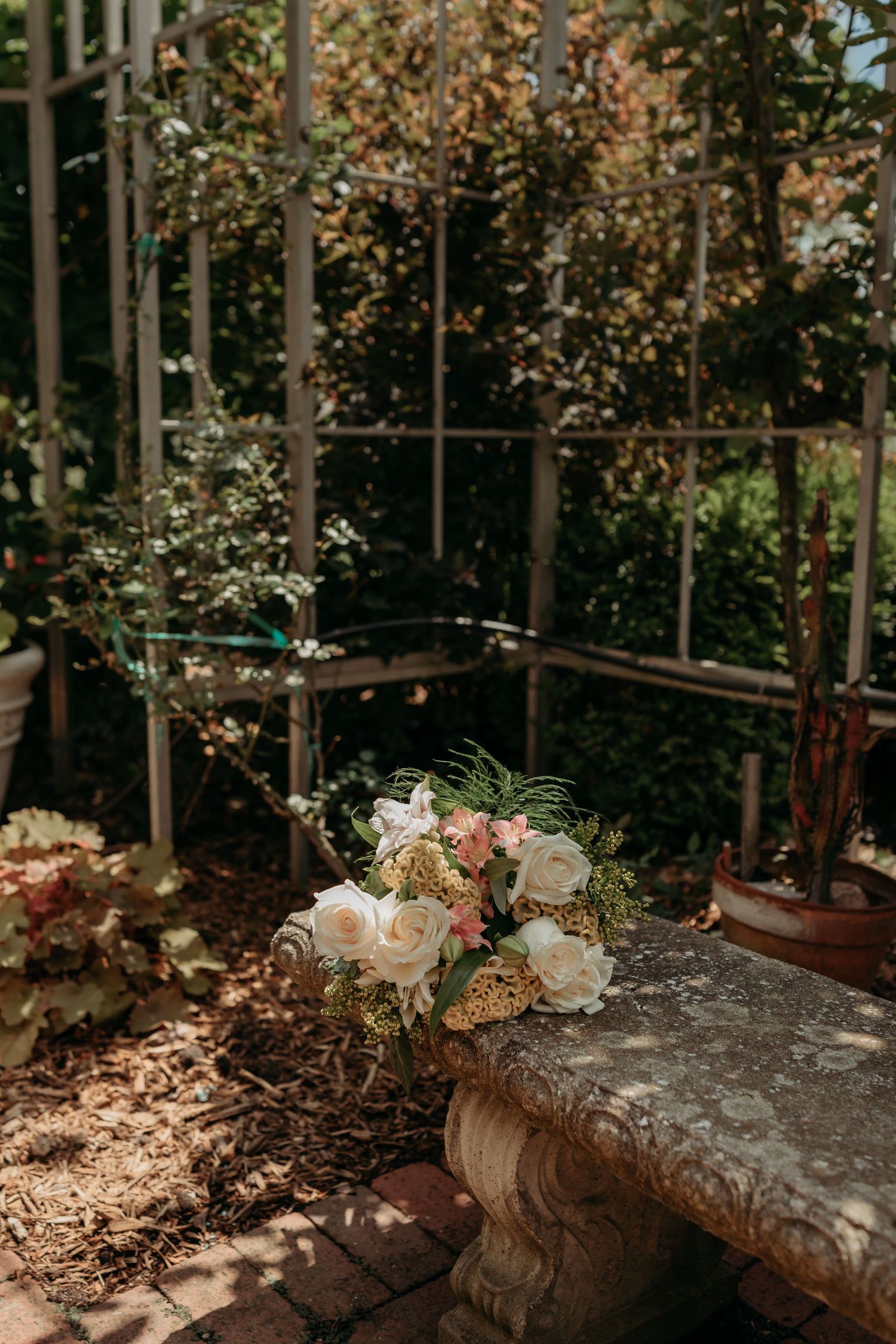A delicate floral bouquet with roses and greenery sits on a stone ledge in a greenhouse with dappled sunlight.