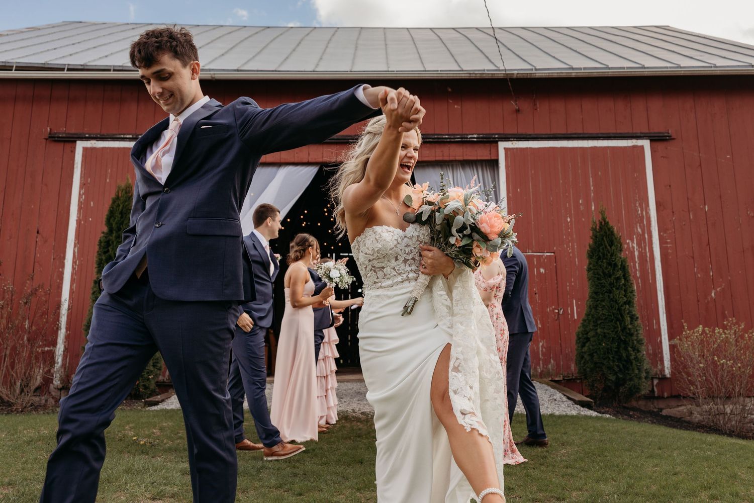 A joyful celebration on a dance floor in front of a red barn with bridesmaids in pink dresses looking on.