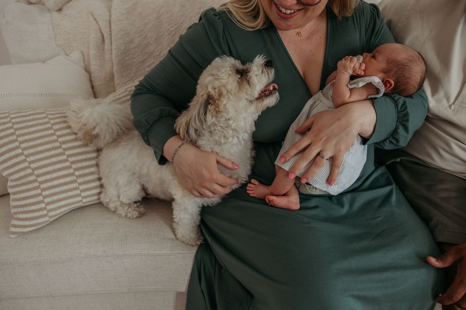 Small white dog sits next to person holding newborn baby on cozy striped couch in green dress and sweater.