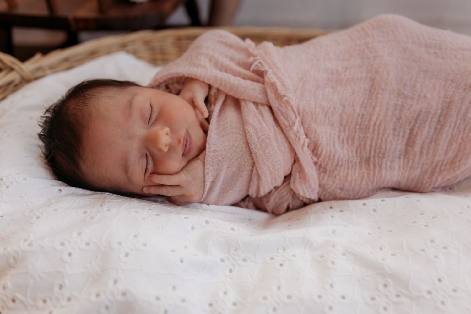 A sleeping baby wearing soft pink lies peacefully on white bedding in a dimly lit room.
