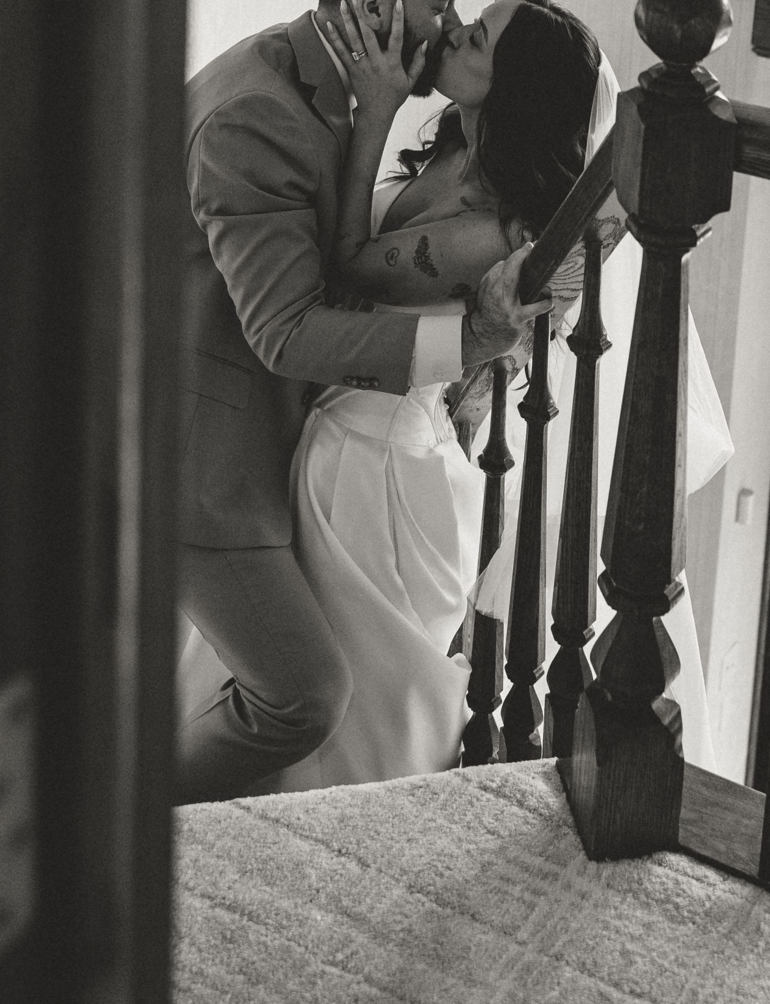 Black and white photo of a couple sharing a romantic kiss on a staircase, the woman in a white dress.