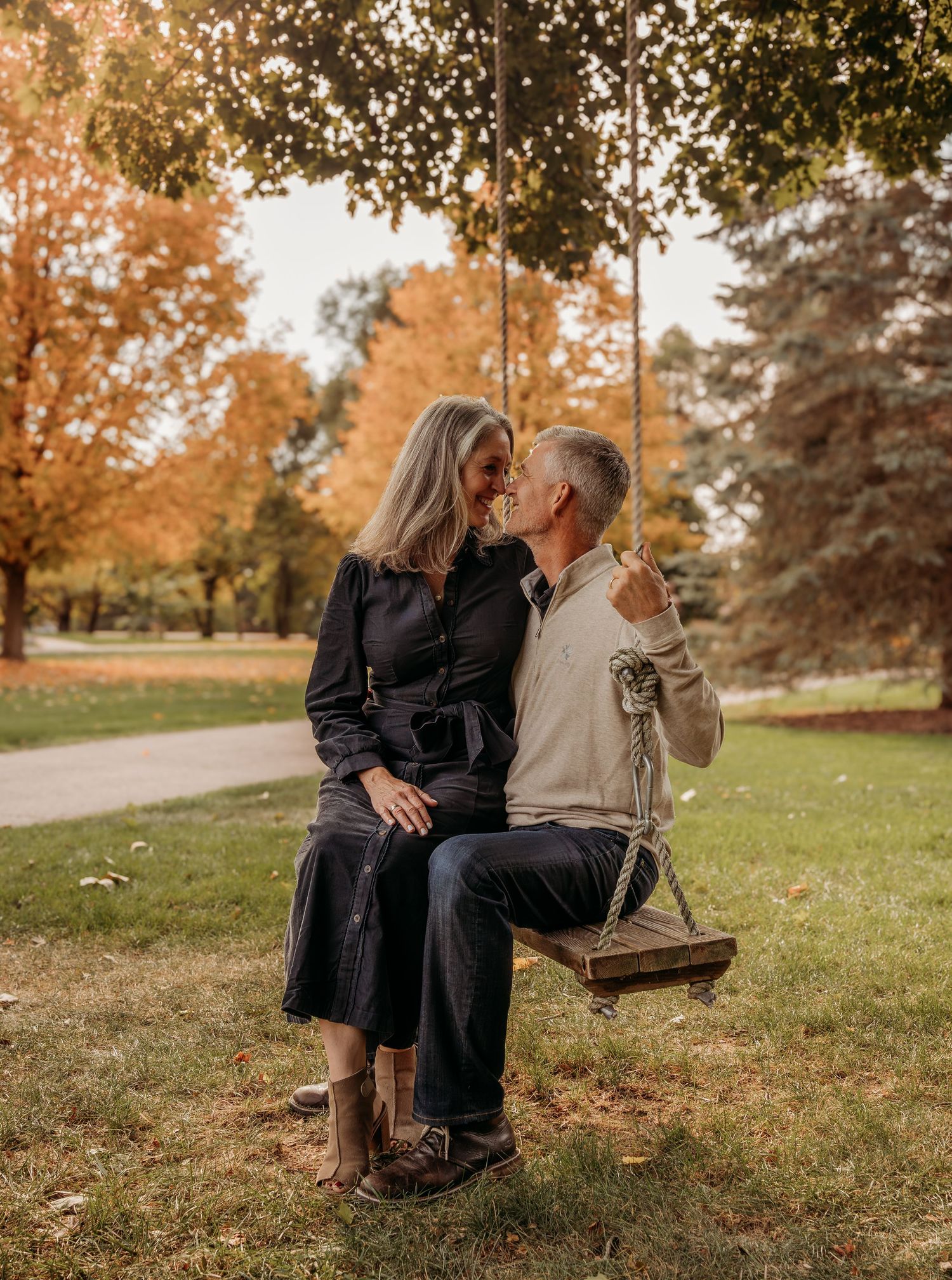 A couple sits together on a wooden bench surrounded by autumn trees with golden foliage in a park setting.