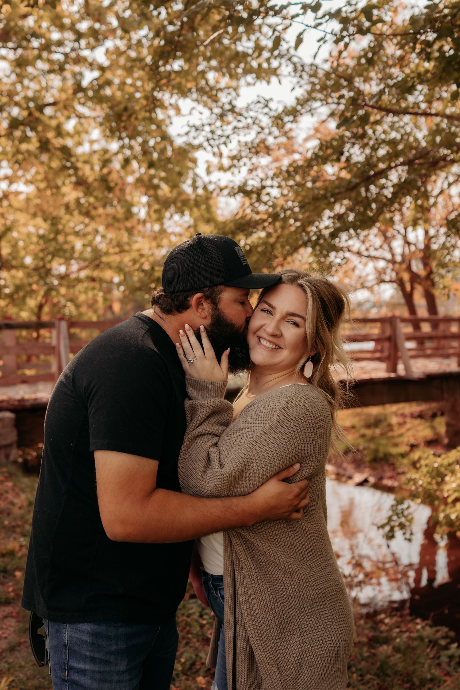 Couple sharing an intimate moment outdoors during autumn with fall foliage and a creek in the background.