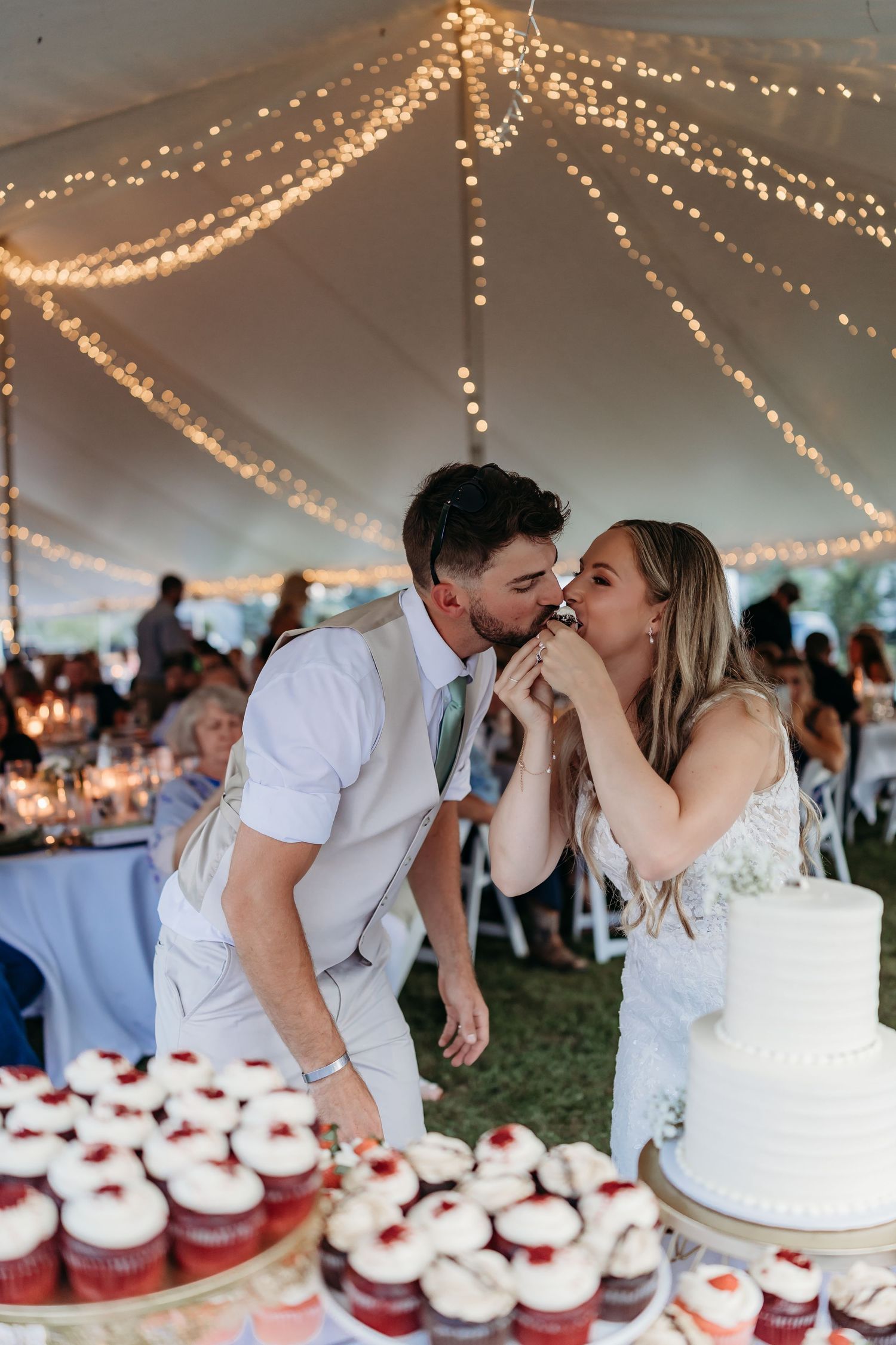 A romantic sequence of newlyweds sharing a sweet kiss behind their wedding cake and cupcakes under a twinkling tent canopy.