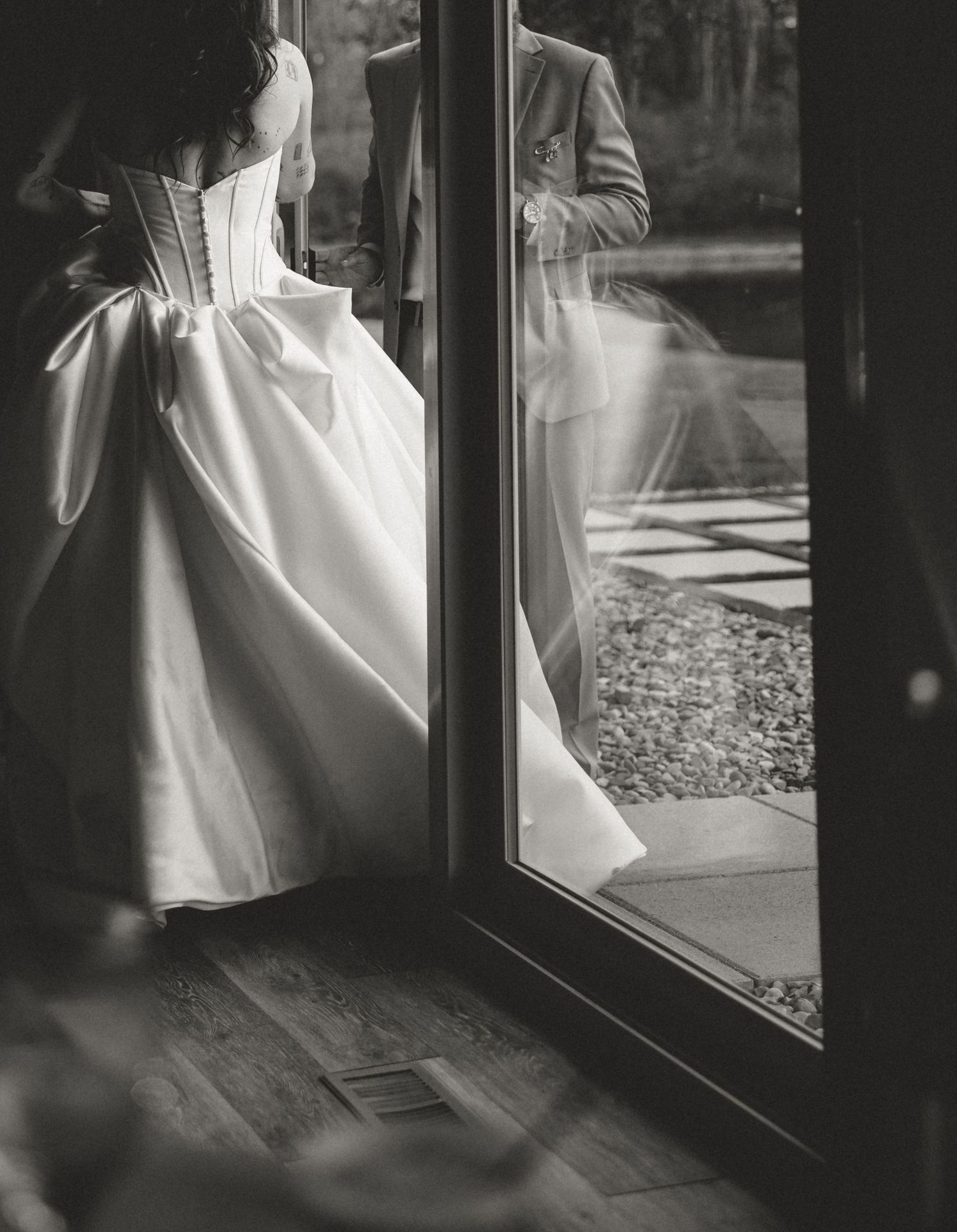 Black and white photo of a bride in a white corset gown standing by glass doors with groom reflection.