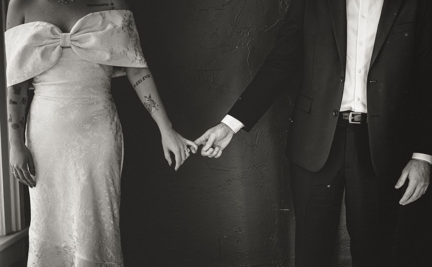 A black and white vintage photograph showing a couple holding hands, with the woman wearing a light dress and bow.