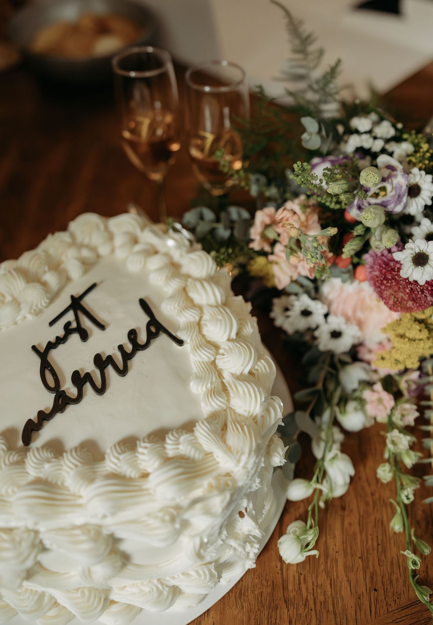 White wedding cake with 'just married' topper beside a colorful floral arrangement and champagne glasses.
