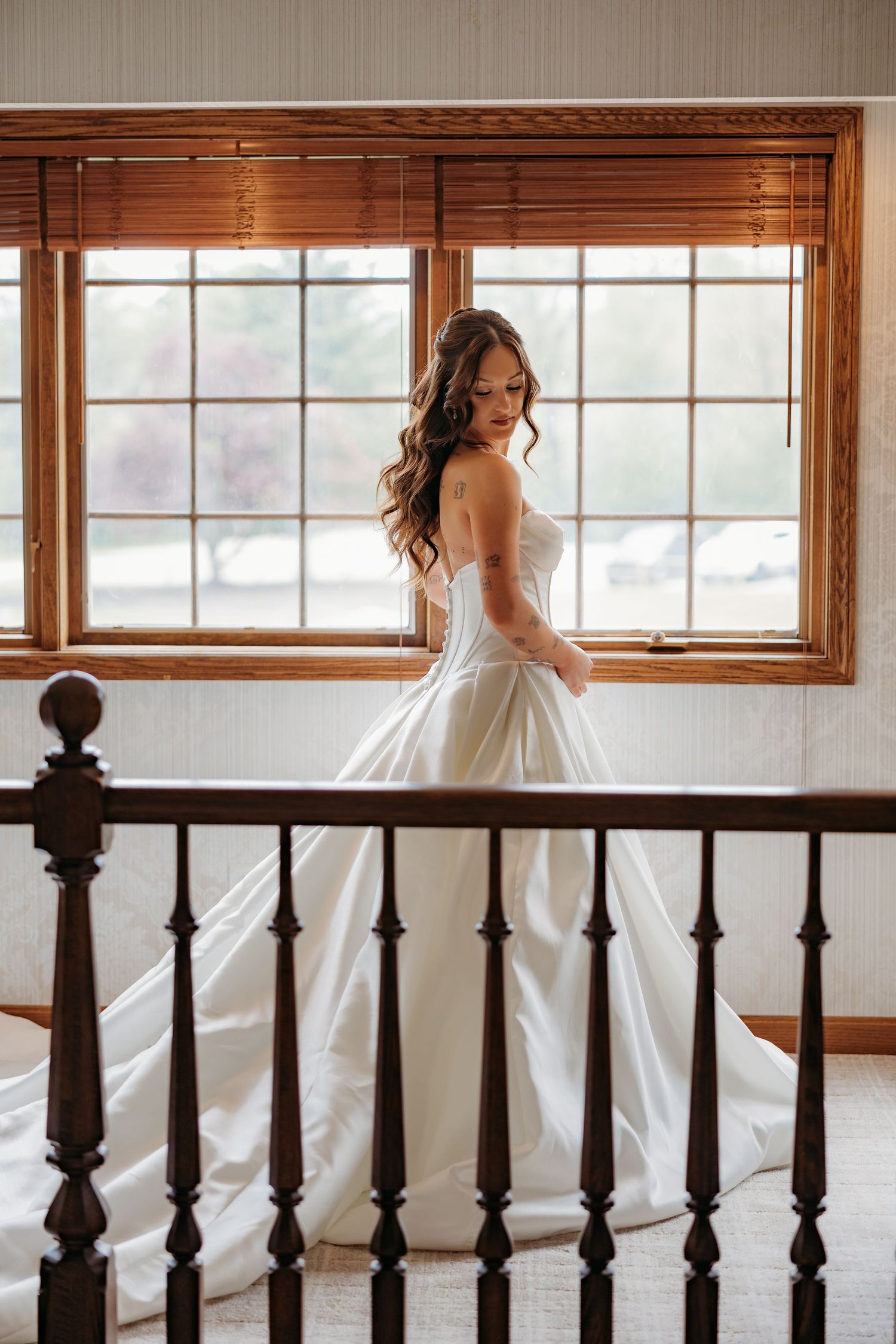 Bride in strapless white ball gown stands by wooden-framed window on staircase landing.