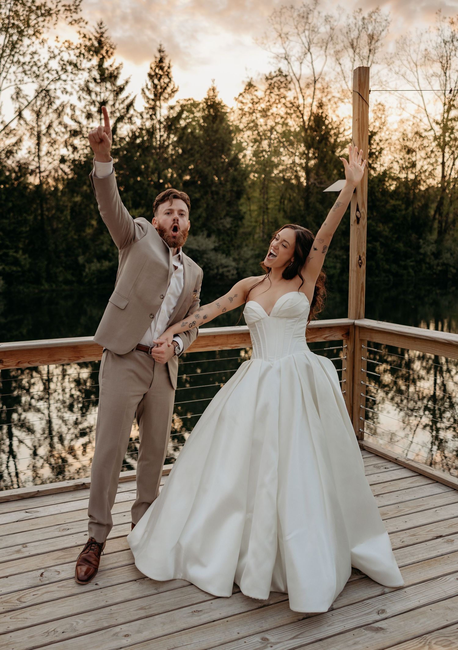 Joyful newlyweds celebrate on a wooden deck at sunset, groom in tan suit and bride in white ballgown, arms raised.