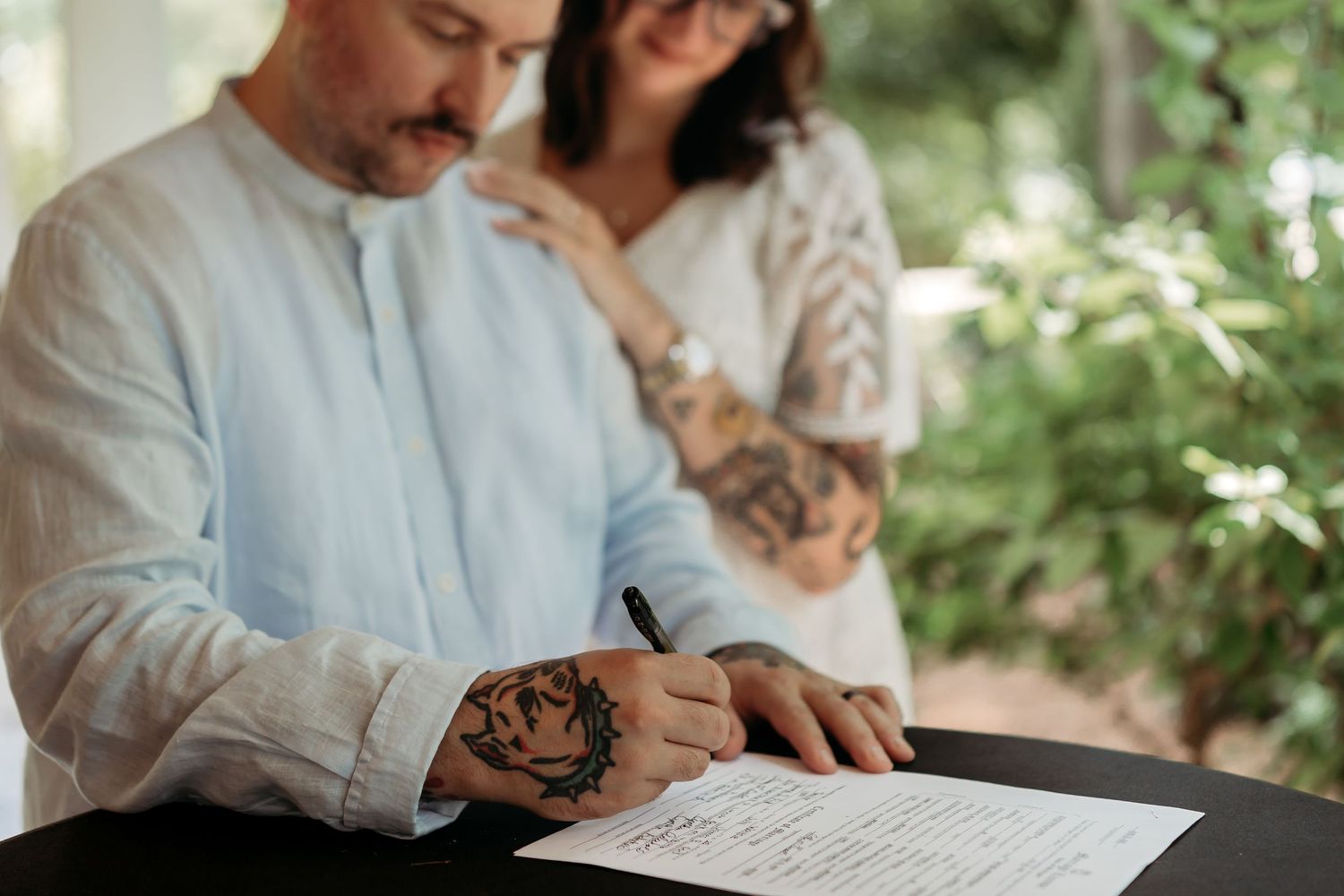 A couple signs documents at an outdoor table surrounded by lush greenery during a ceremony.