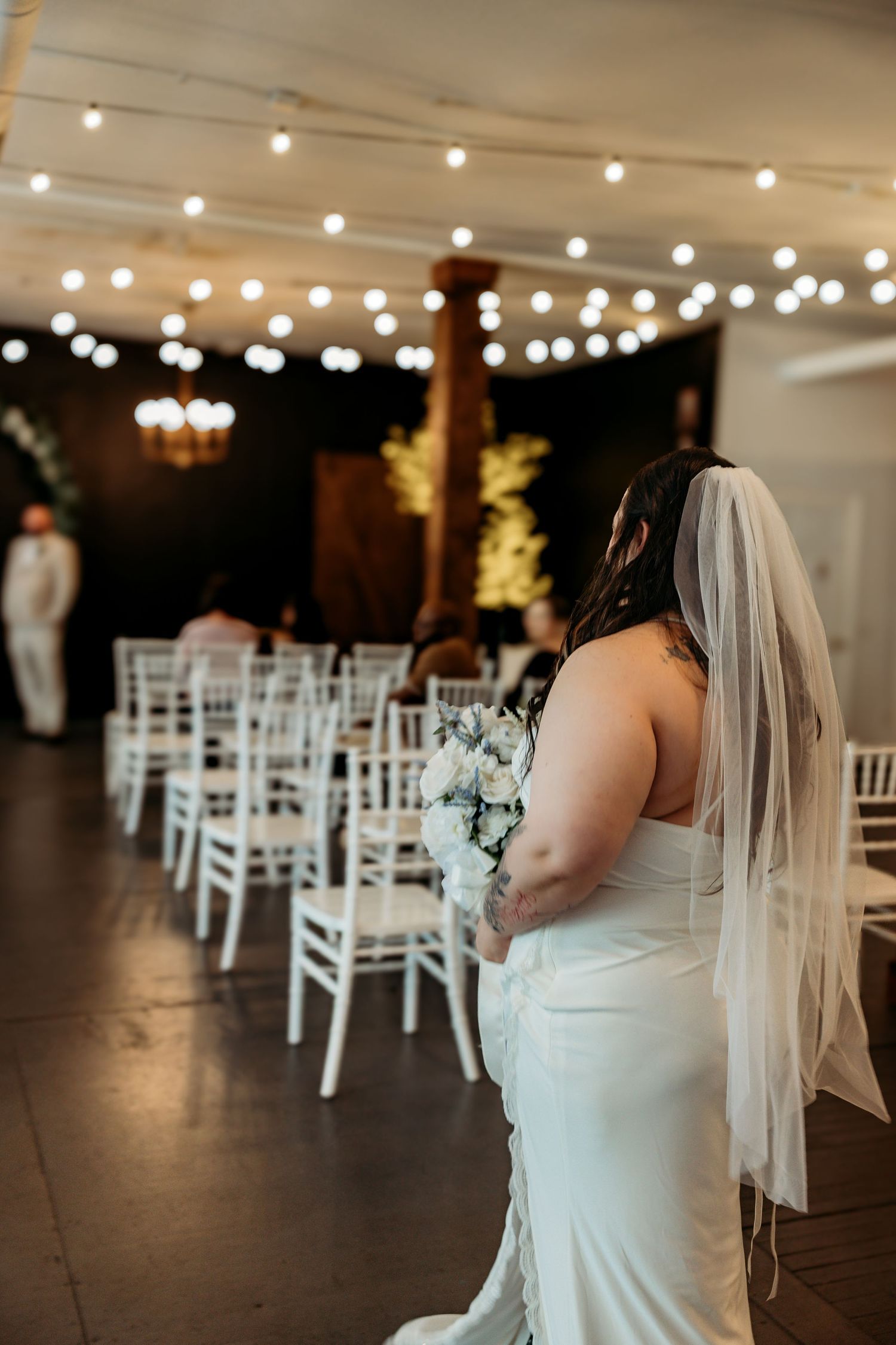 Wedding ceremony setup with white chiavari chairs and string lights creating an elegant indoor venue atmosphere.