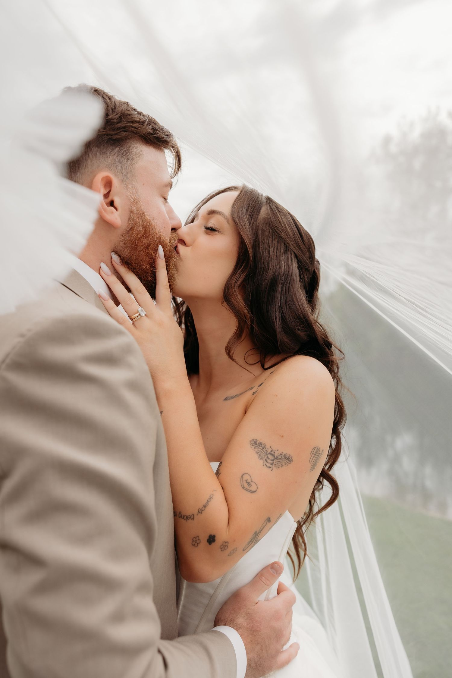 Bride and groom sharing a romantic kiss under a white veil, bride wearing a strapless gown with tattoos and engagement ring.