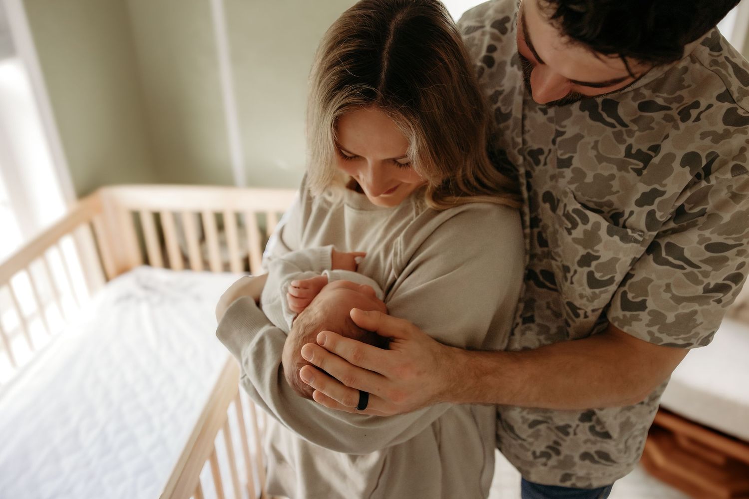 Military service member and partner hold newborn baby near wooden crib in soft natural light.