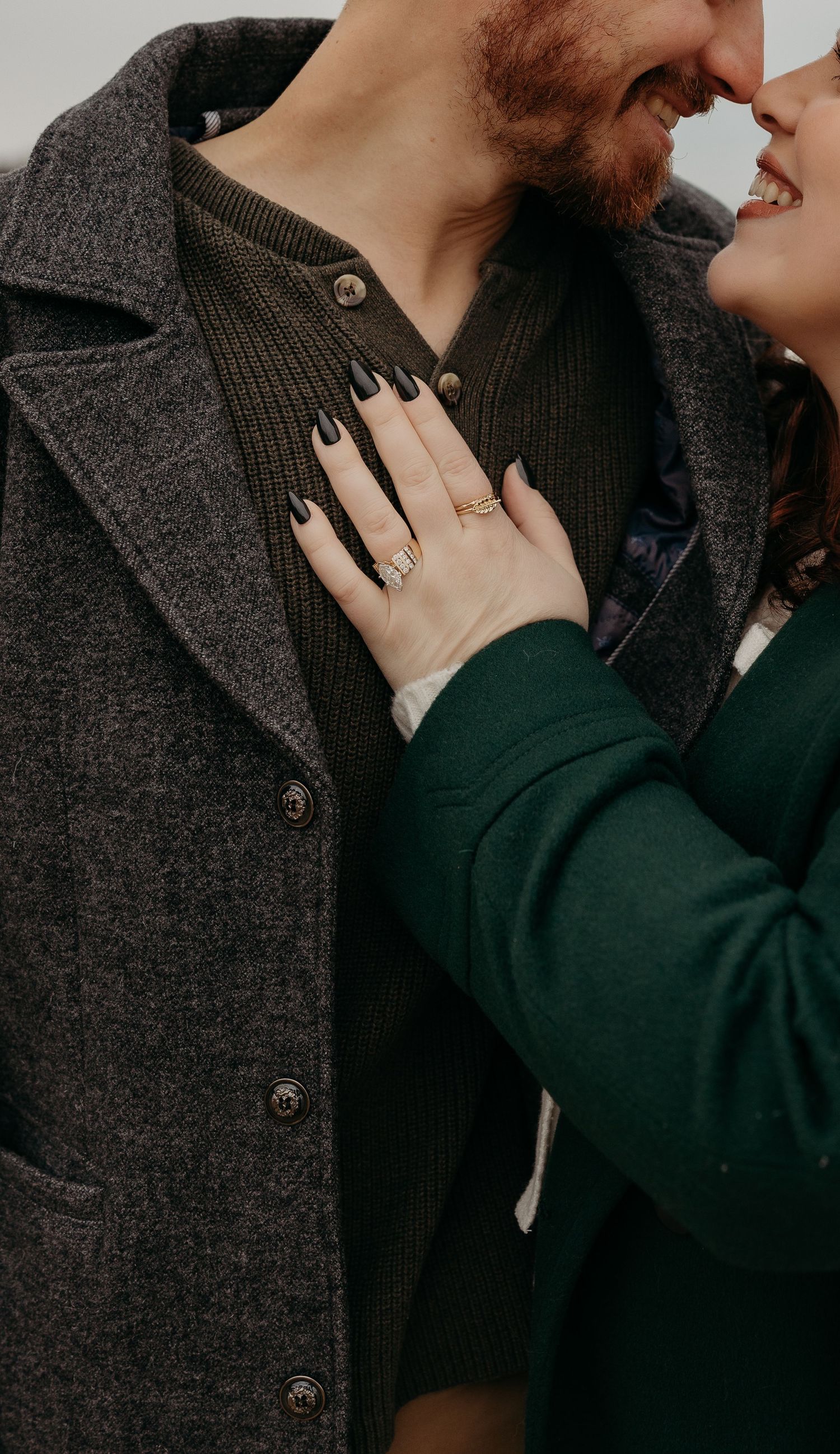 Close up of couple embracing while wearing dark winter coats, with an engagement ring visible.