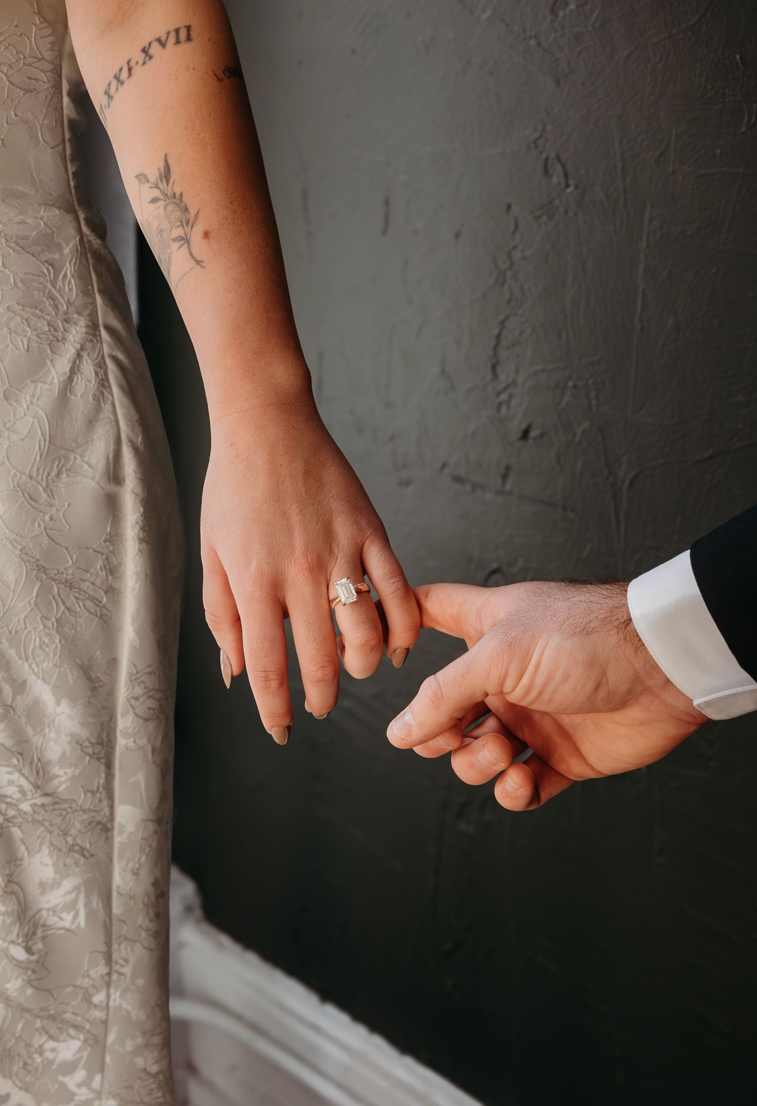 Close-up view of a couple holding hands with a wedding ring visible against a dark gray background.