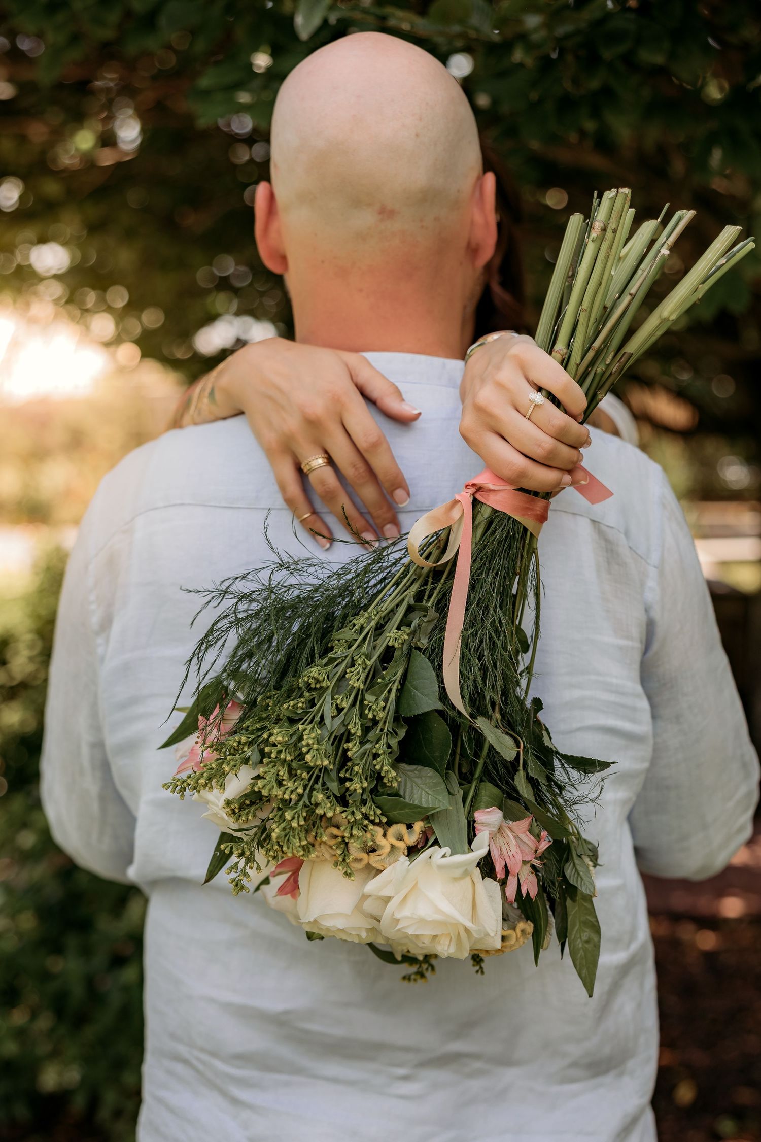 Back view of someone in white shirt holding bouquet of fresh flowers and greenery against sunlit garden background.