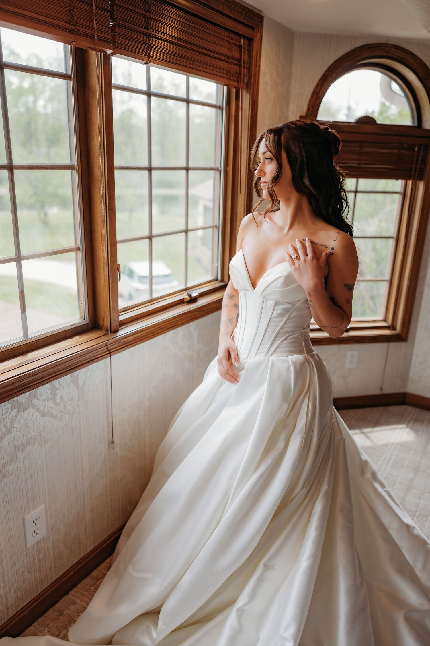 Bride in a white strapless ball gown gazes out wooden-framed windows in a softly lit room.