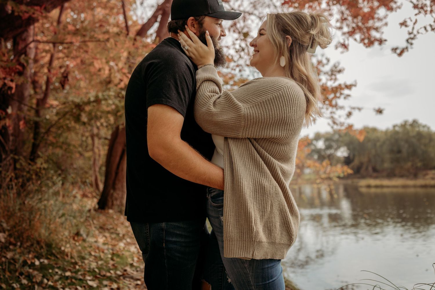 Romantic couple embraces by a serene lake during autumn with orange foliage reflecting in the water.