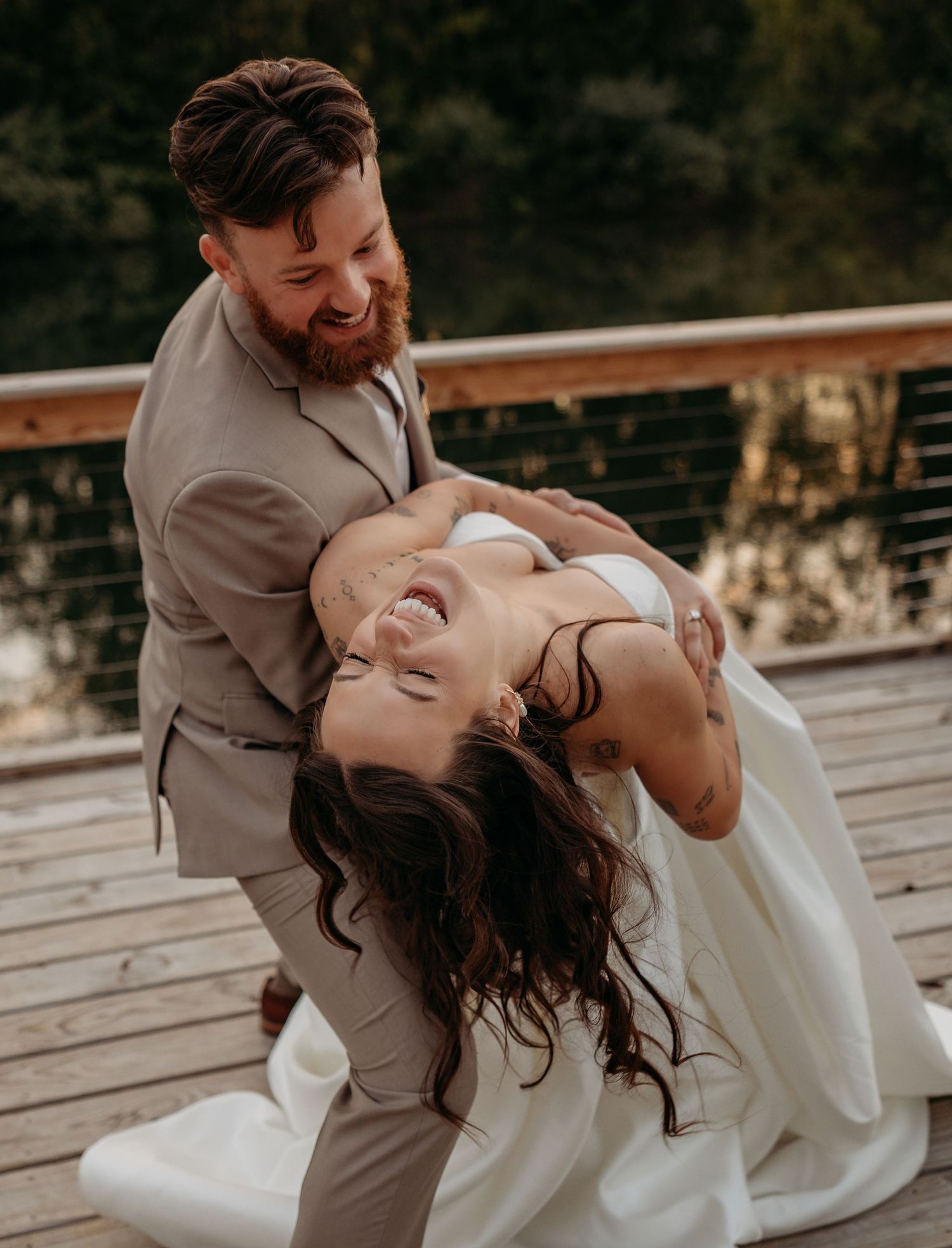 A joyful bride laughs as her bearded groom dips her on a wooden deck during their outdoor wedding celebration.