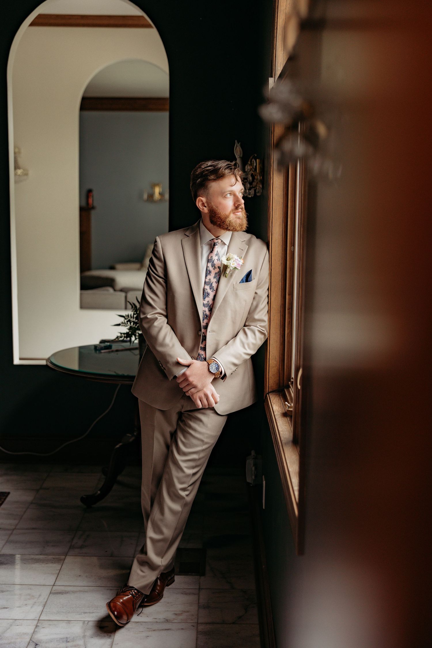 Groom in tan suit with floral tie leans against window, gazing out in elegant interior setting.