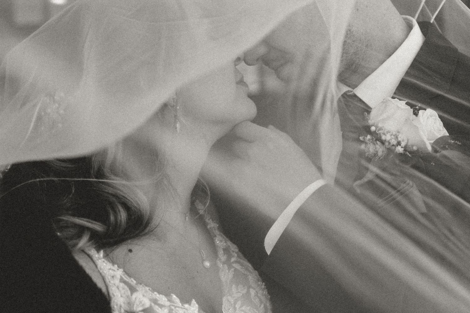A tender romantic kiss between a couple in wedding attire, captured through delicate white tulle veil in black and white.