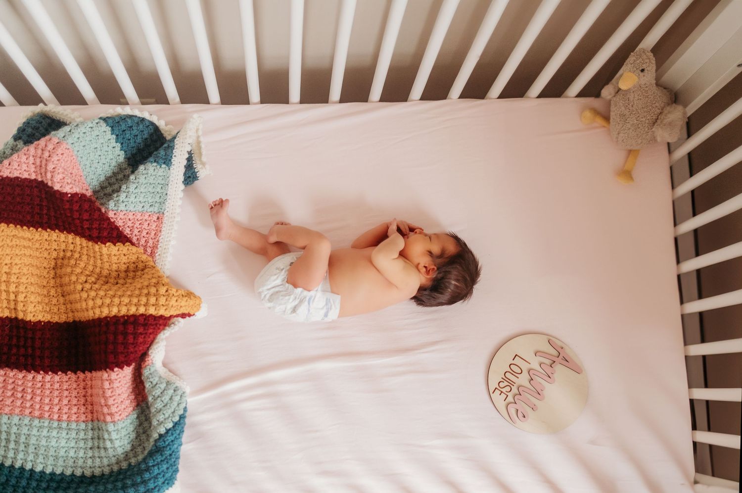 A baby wearing colorful striped knitwear lies on white bedding with multiple matching outfits arranged nearby.