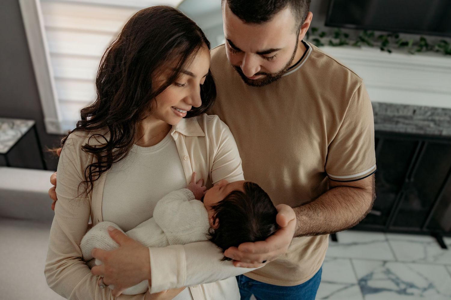 A couple in neutral colored sweaters lovingly holds and gazes at their dark-haired baby in a modern indoor setting.