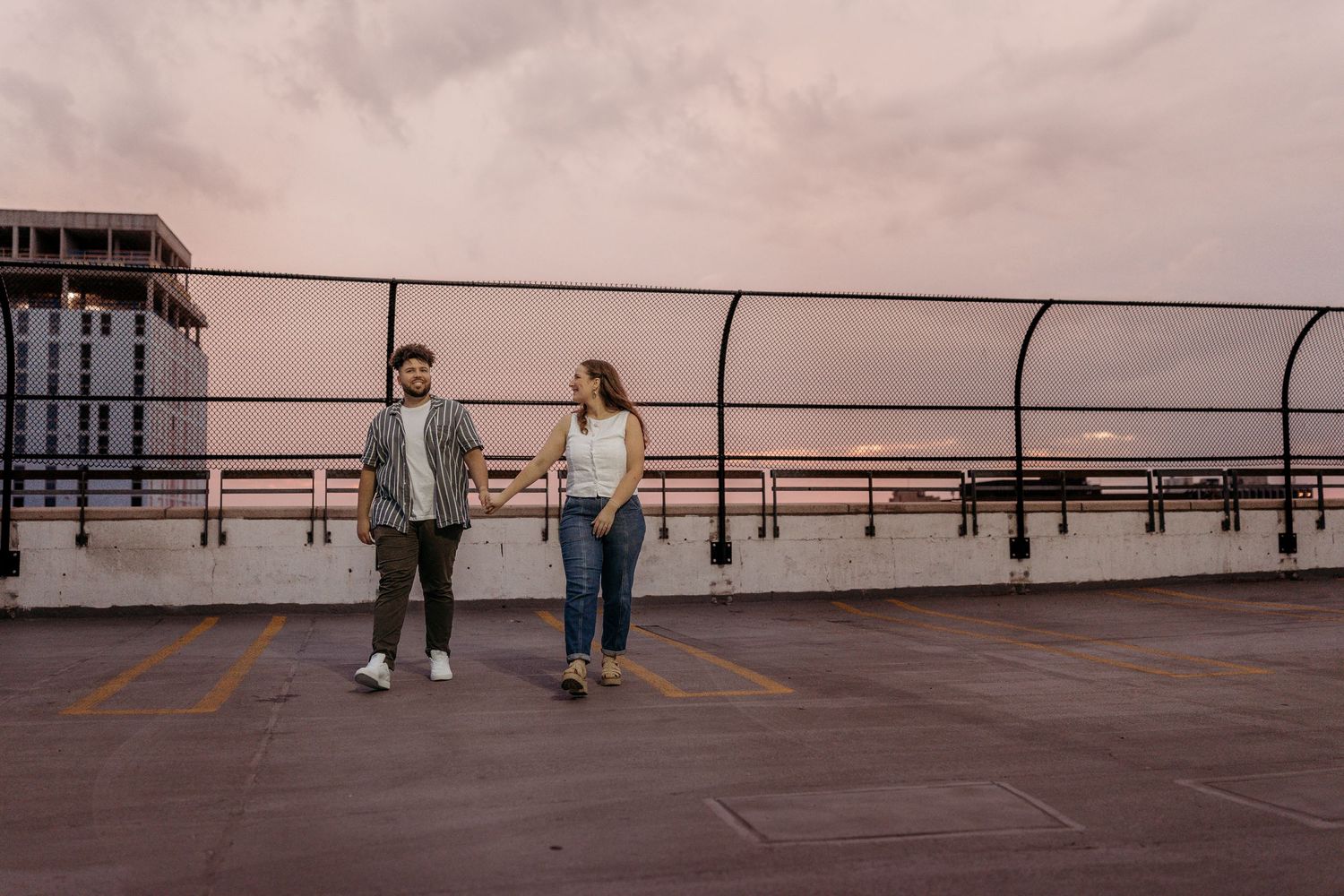 Two people walk together on a rooftop deck with railings at sunset against a moody pink sky.