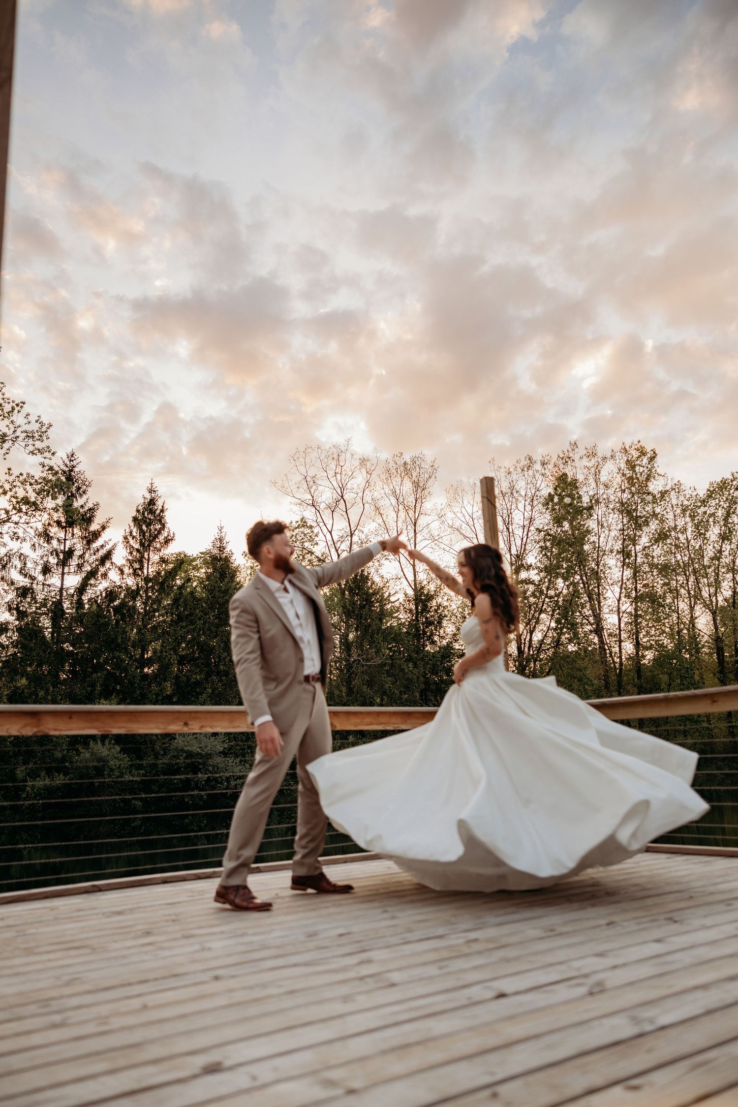 Bride and groom dancing on outdoor deck at sunset with flowing white dress twirling against dramatic sky.