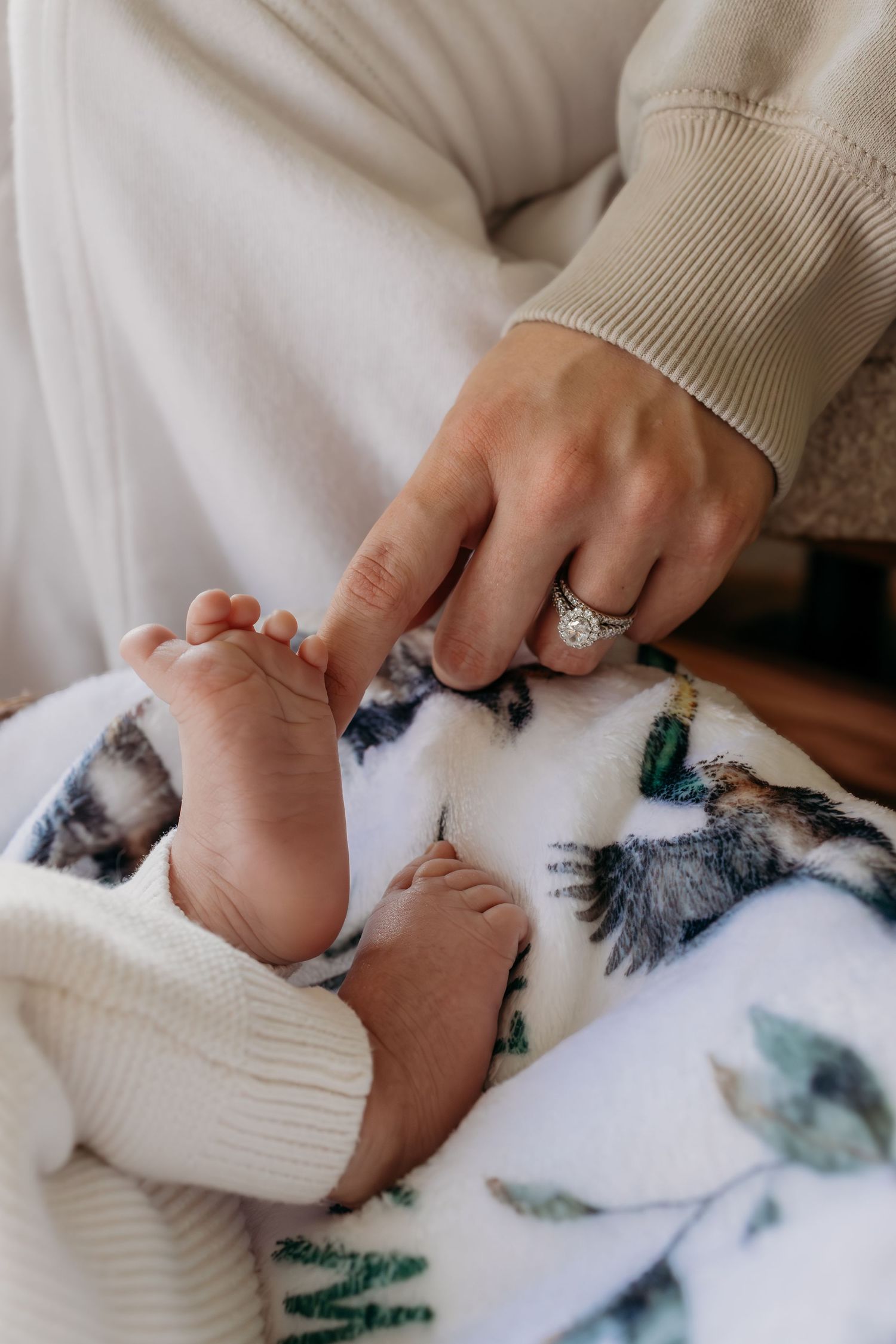 A gentle hand wearing an engagement ring touches tiny baby feet on a patterned white blanket.