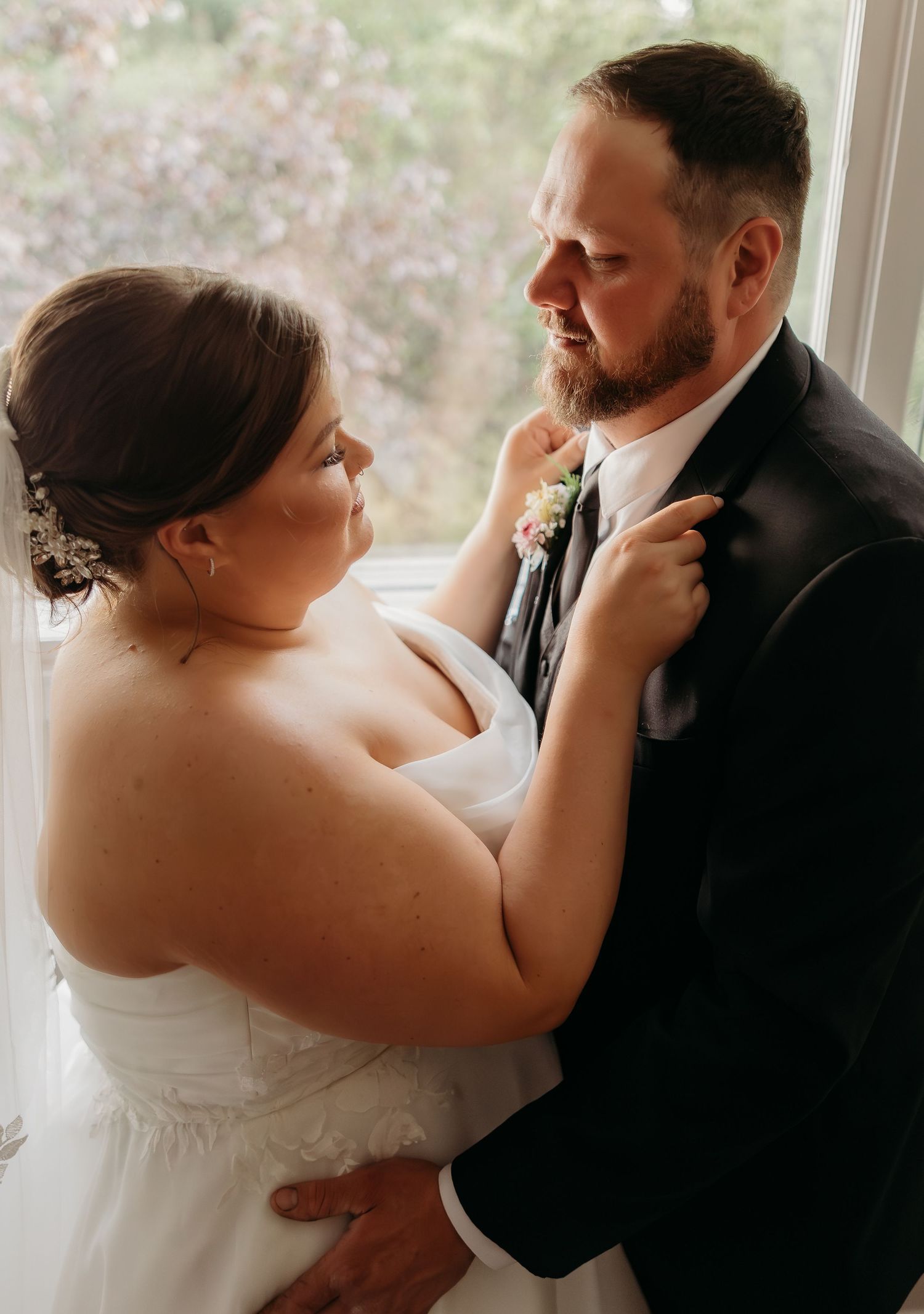 Bride in white gown adjusts groom's boutonniere near window in intimate wedding portrait.