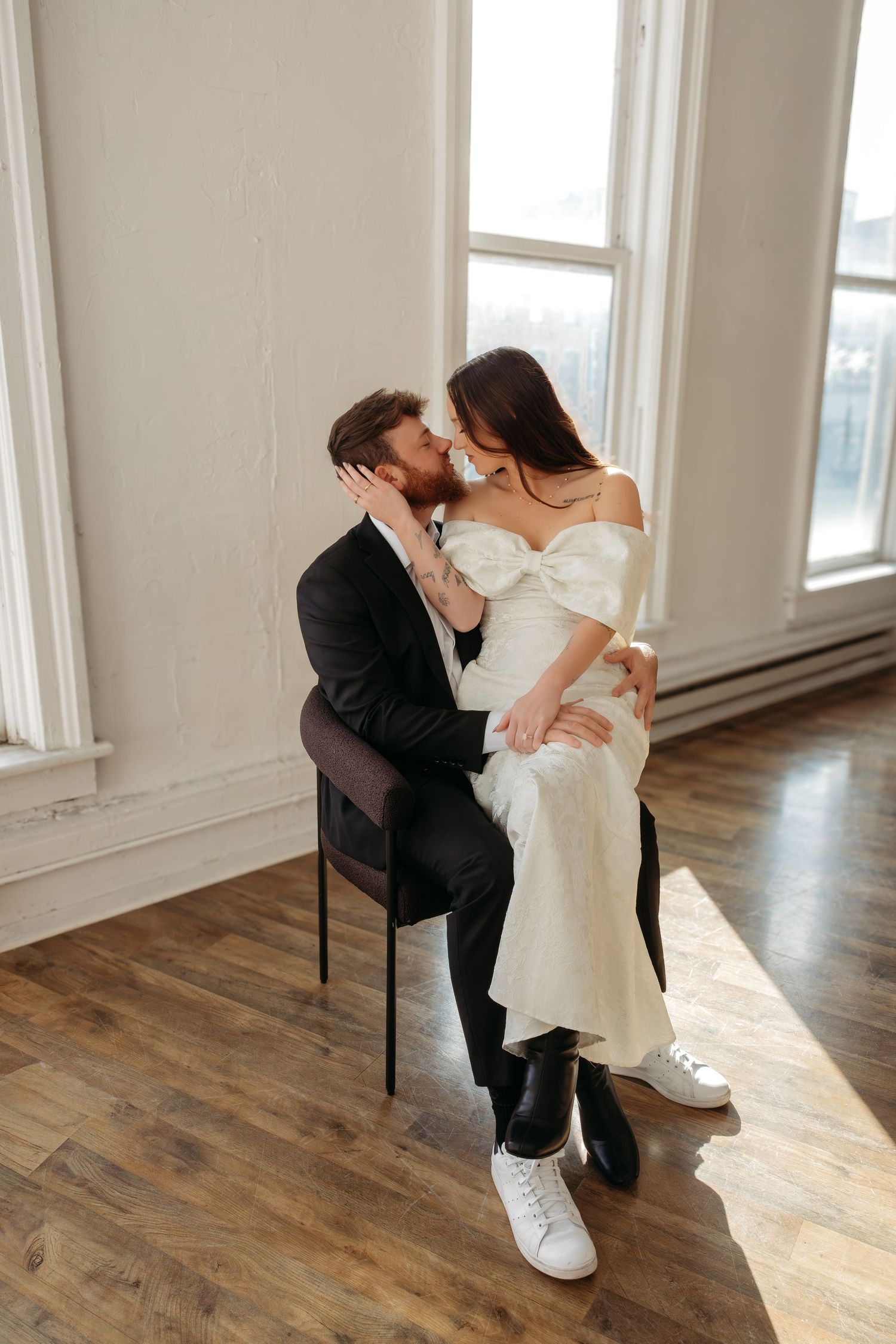 Romantic wedding couple embraces while seated on wooden chair by bright window in minimalist room with hardwood floors.