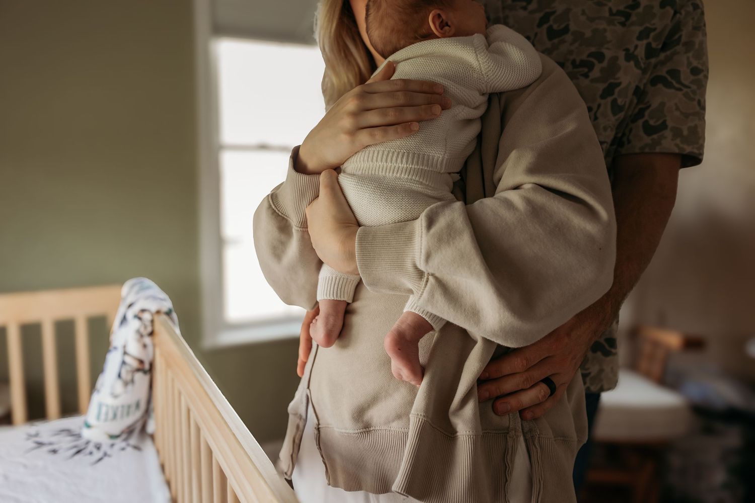 A parent holds a small baby wearing neutral colored clothes next to a wooden crib in a softly lit room.