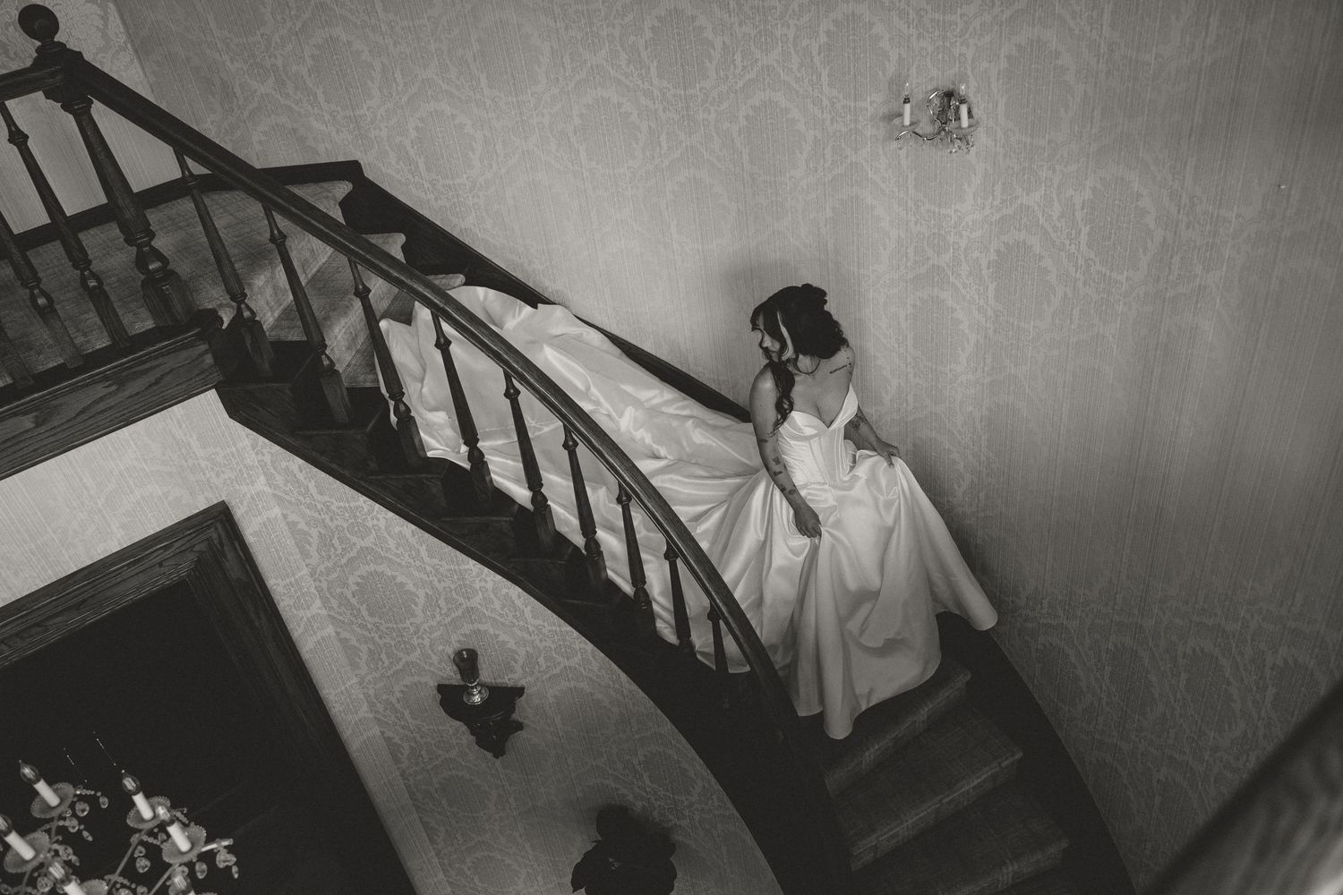 Bride in white gown descending elegant staircase in black and white wedding photo.