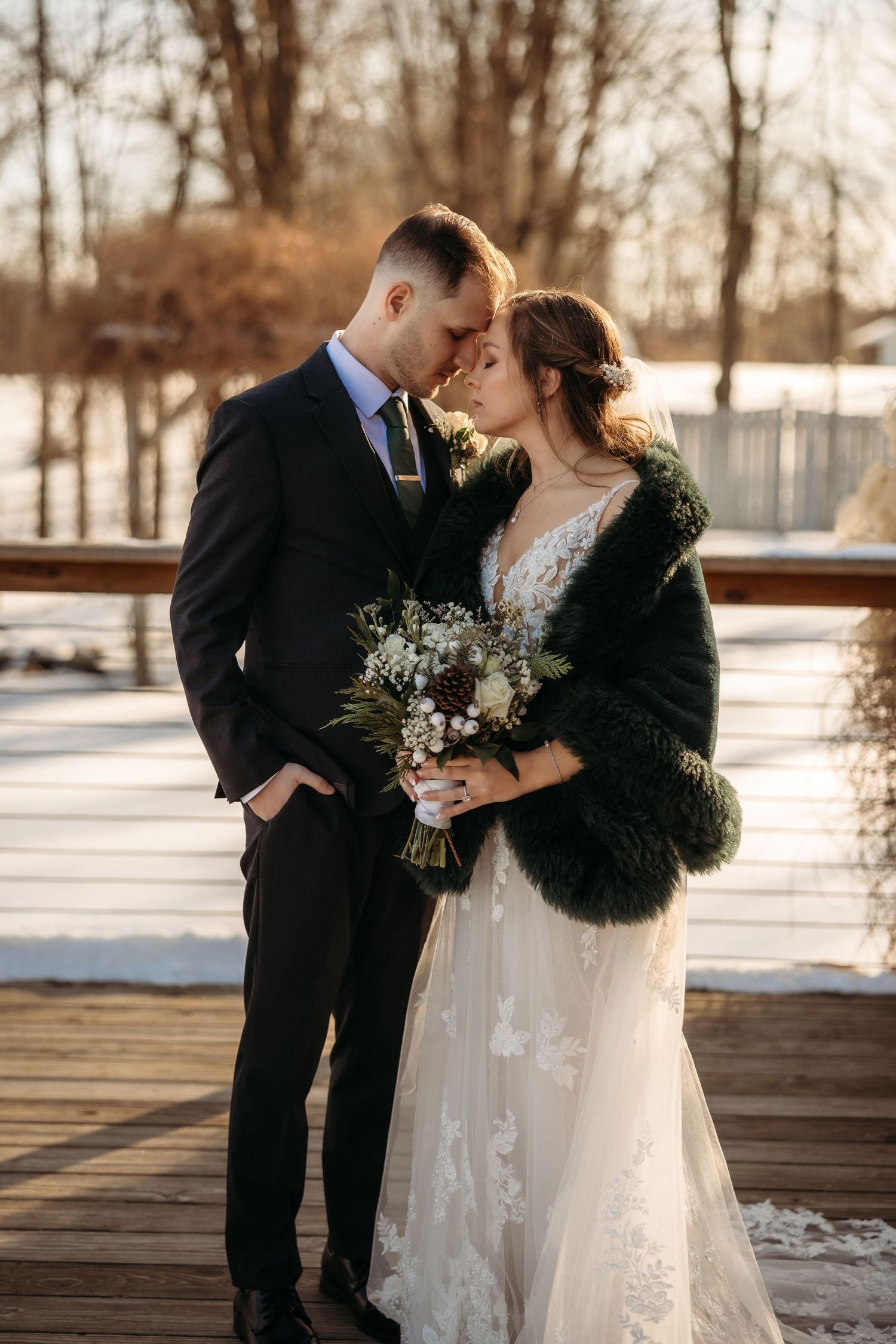 A winter wedding photo of a couple embracing on a wooden dock as sunlight glows behind them, the bride holding a delicate bouquet.