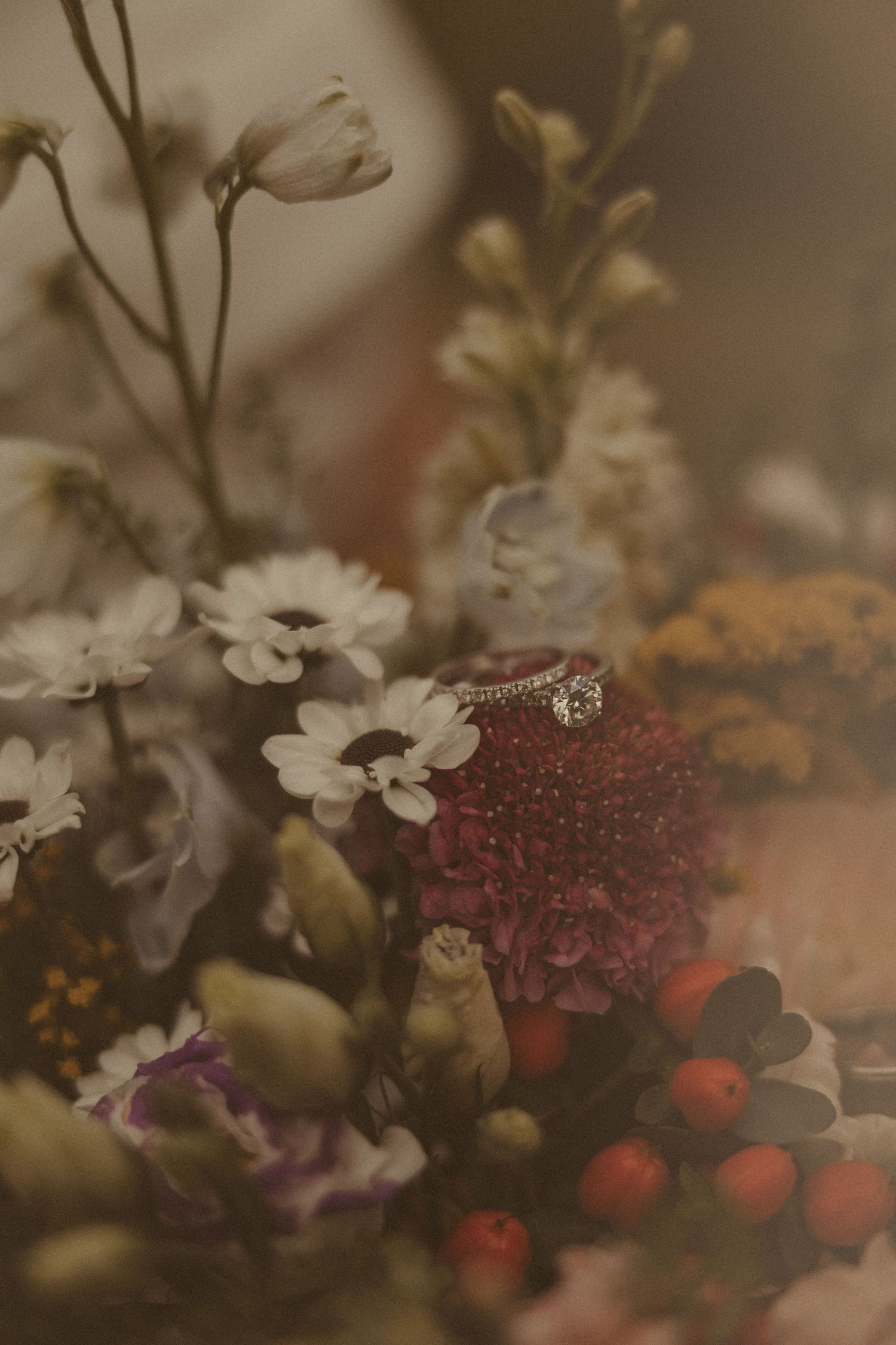 Close-up of a diamond engagement ring nestled among colorful wedding flowers including daisies and red berries.