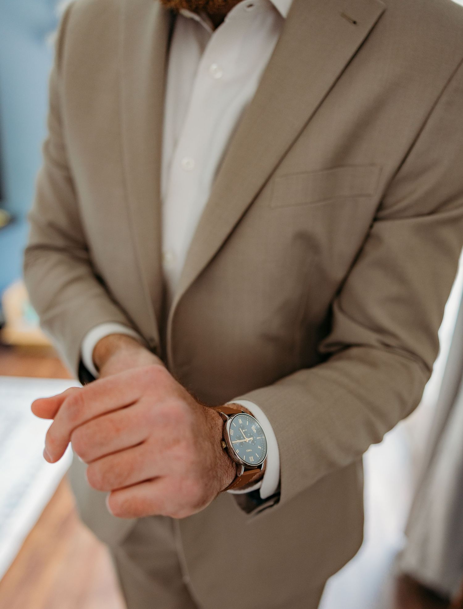Man in beige suit adjusting his watch, wearing a classic brown leather timepiece with a dark dial.