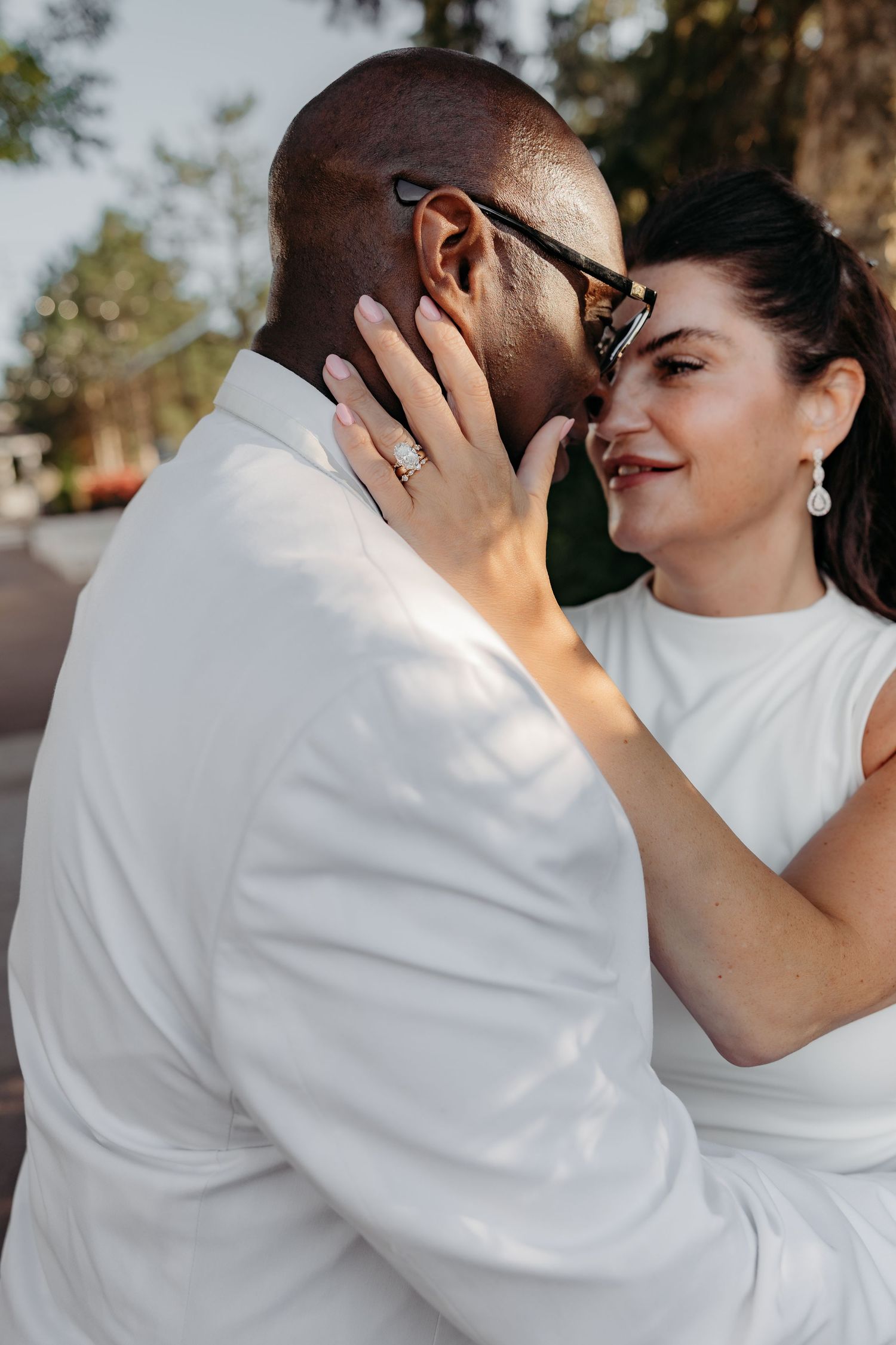 A romantic couple in white outfits embrace intimately during an outdoor photoshoot in warm sunlight.