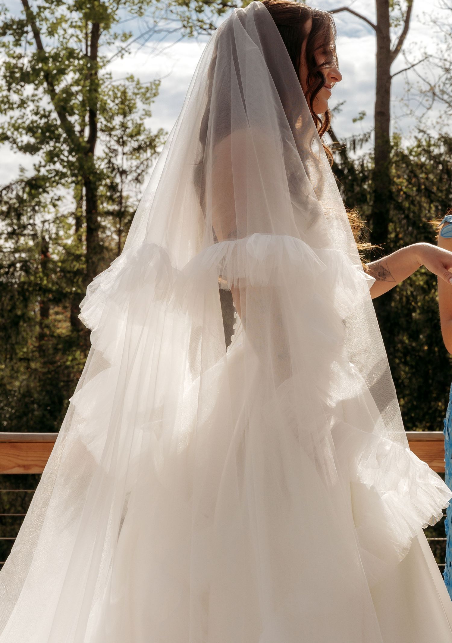 Bride wearing a layered tulle wedding gown and long veil outdoors surrounded by trees.