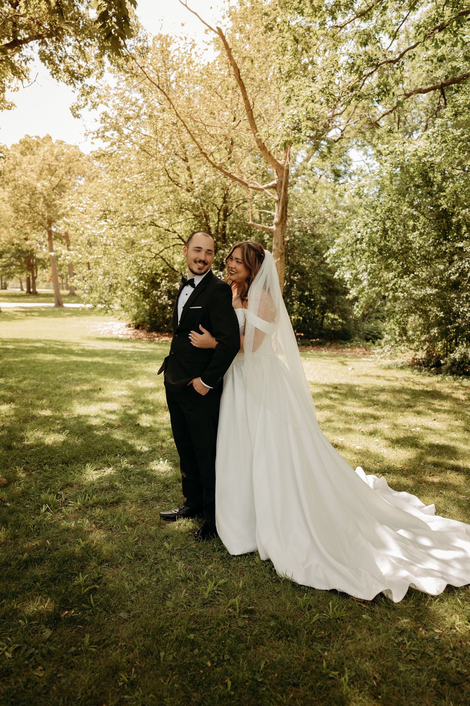 Bride in white gown with veil and groom in black tuxedo embracing outdoors in lush green park setting.