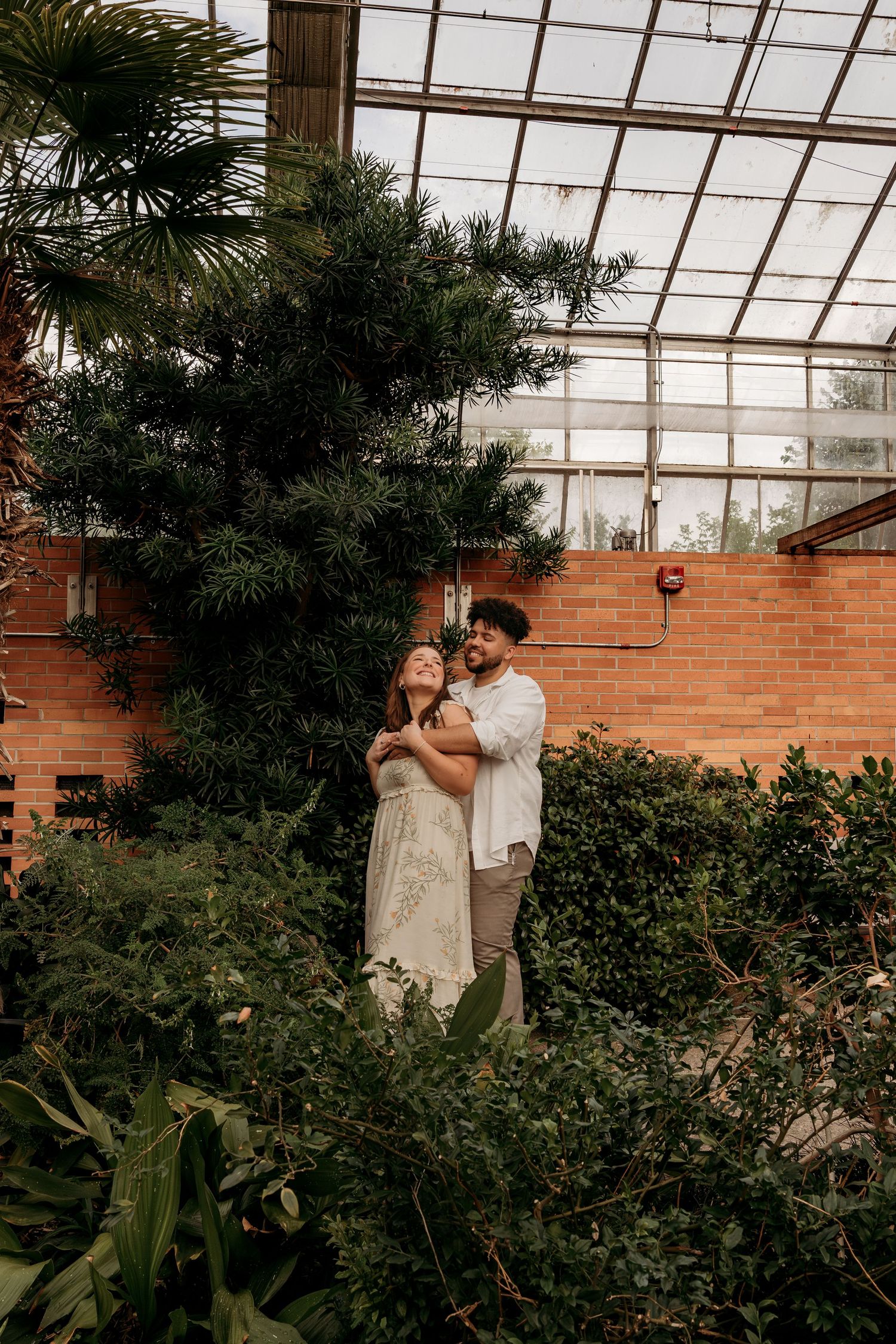 A couple shares a romantic moment in a greenhouse surrounded by lush greenery and brick walls.