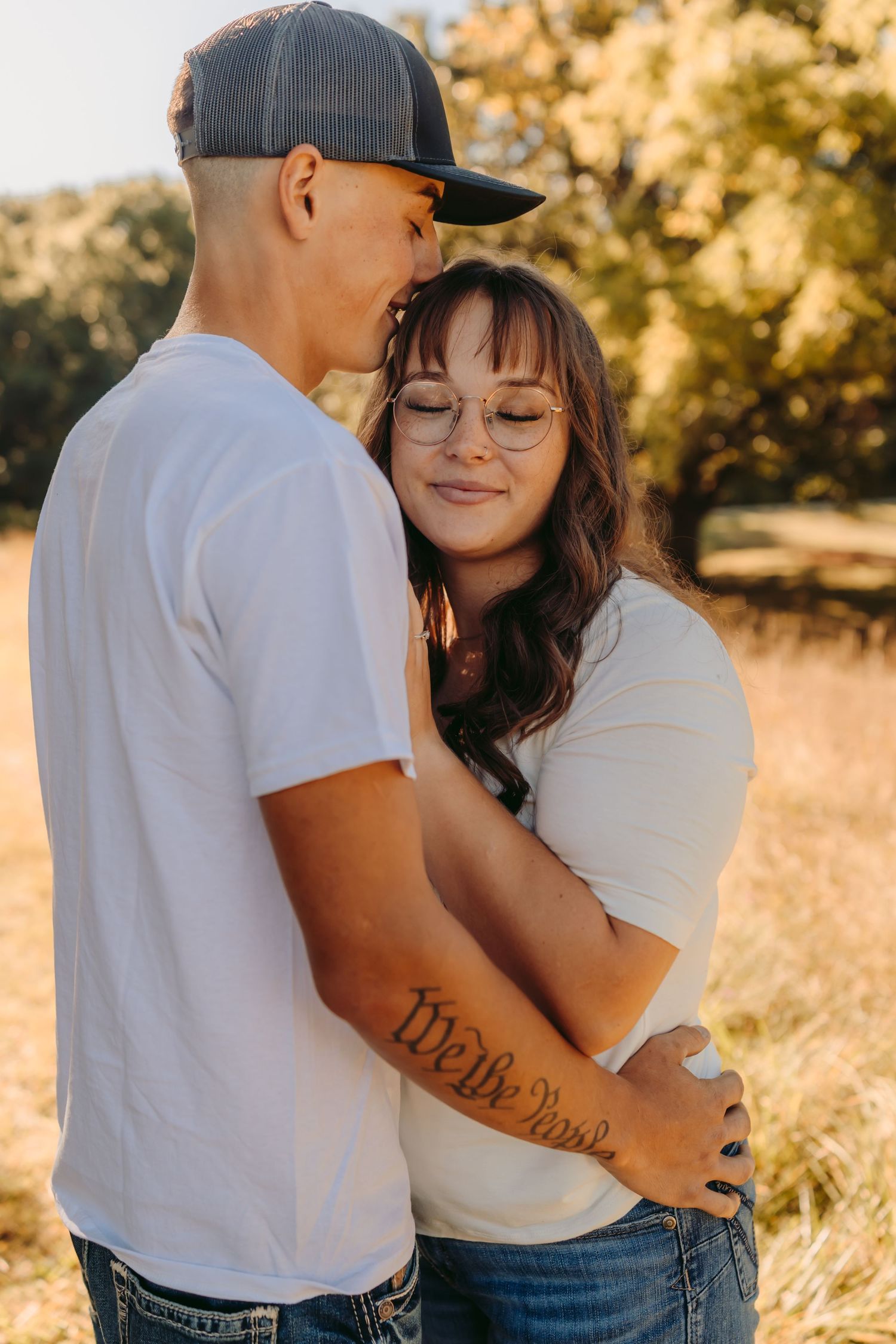 Couple in white shirts and jeans embrace lovingly in a golden sunlit field during sunset.